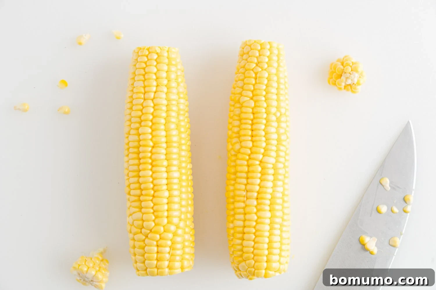 Two ears of corn with the tips trimmed off, next to a sharp knife, illustrating preparation.