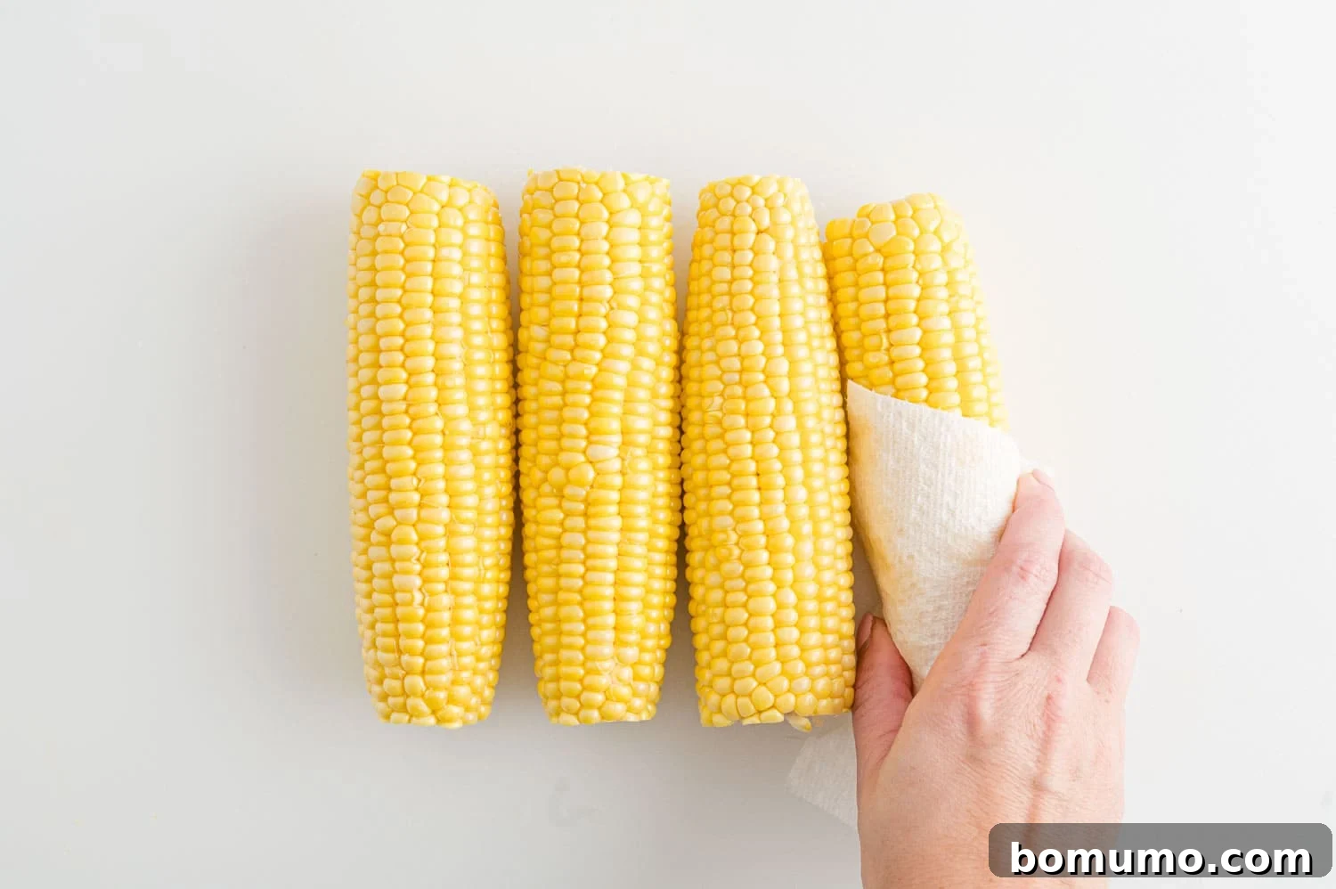 A hand uses a paper towel to pat corn on the cob dry after rinsing, highlighting a crucial prep step.
