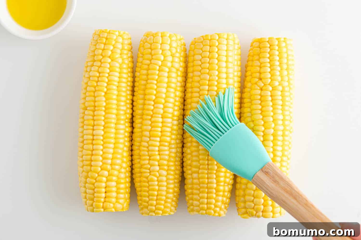 A rubber basting brush is used to brush ears of corn with olive oil, showing the seasoning process.