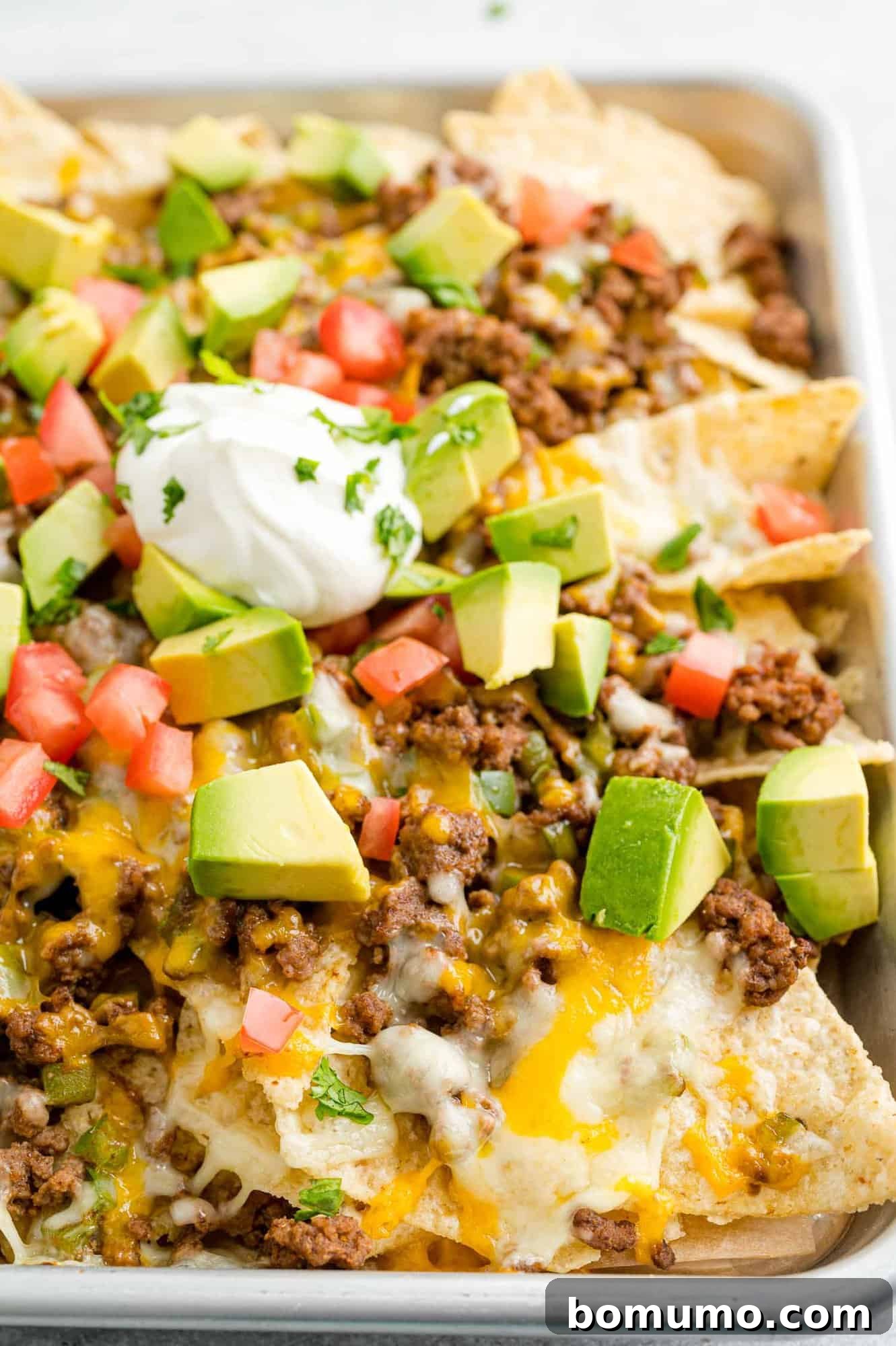 Close up view of a tray of beef nachos topped with cheese, diced tomatoes, avocados, onions, and dollops of sour cream.