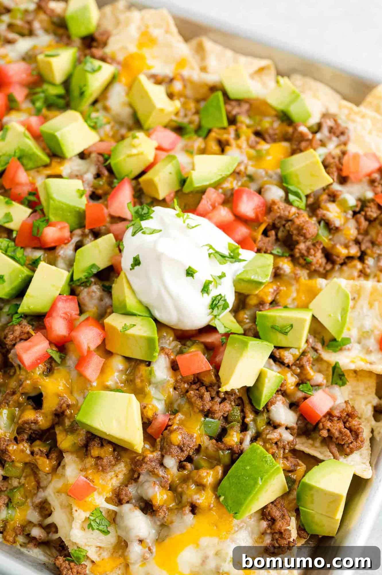 Close up view of a tray of beef nachos topped with cheese, diced tomatoes, avocados, onions, and dollops of sour cream.