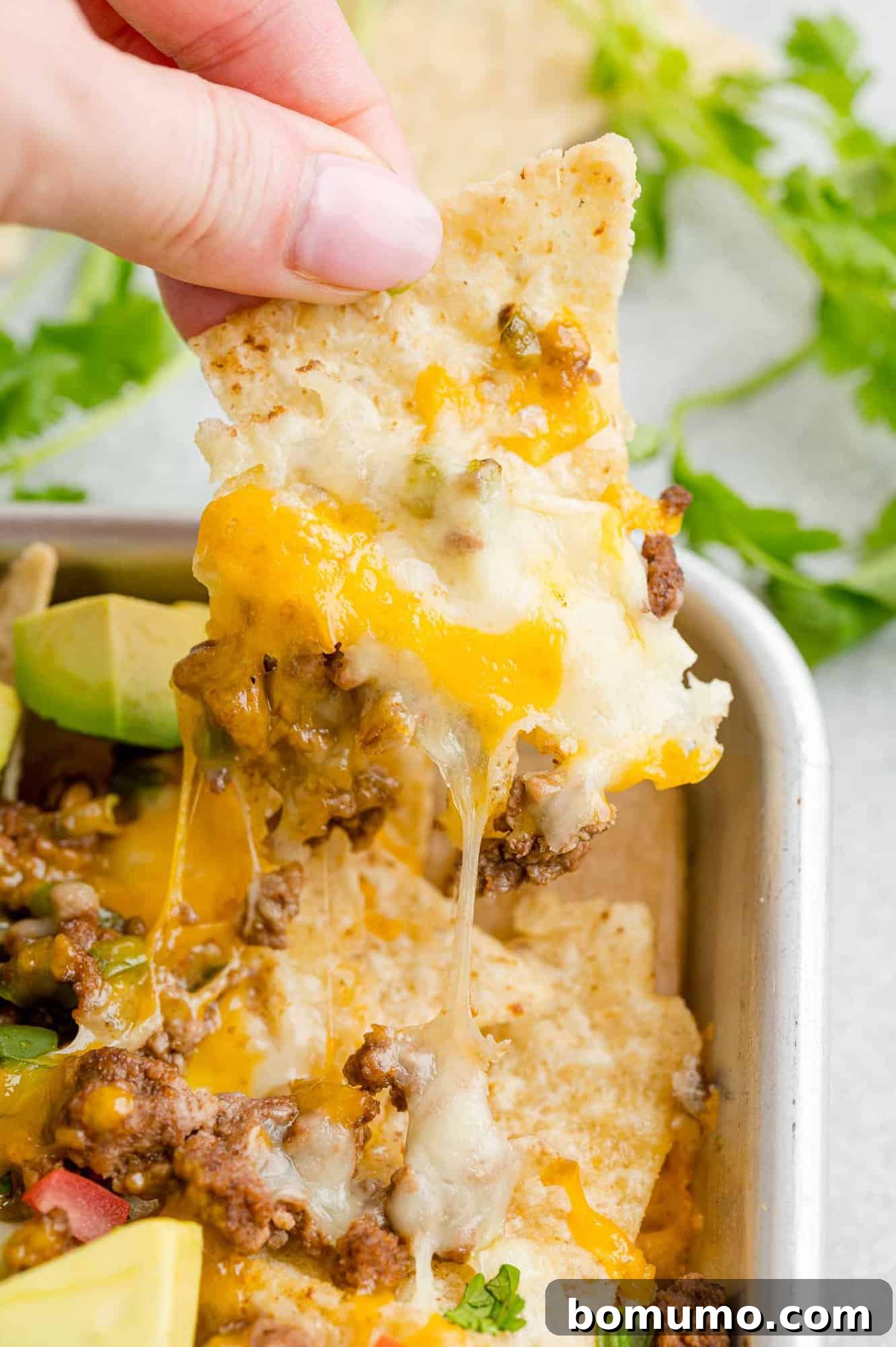 A hand lifts a cheesy tortilla chip from the corner of a pan of beef nachos.