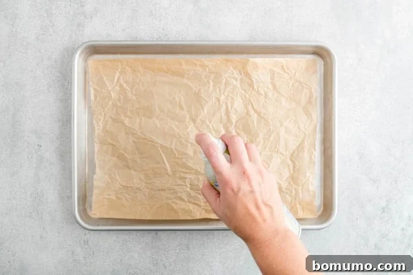 A hand holding a can of cooking spray over a baking sheet lined with brown parchment paper.