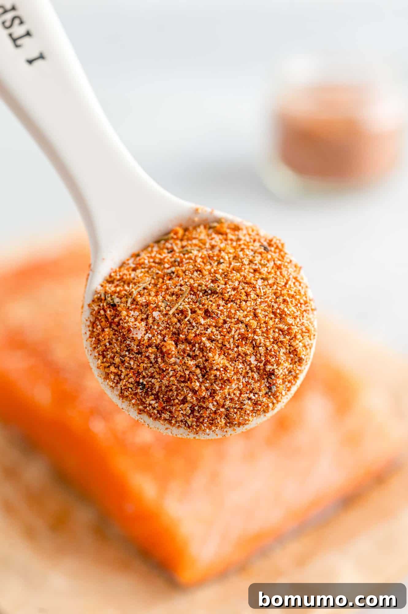 A spoonful of vibrant salmon seasoning is held elegantly in front of a fresh, raw salmon fillet, ready to be seasoned. 