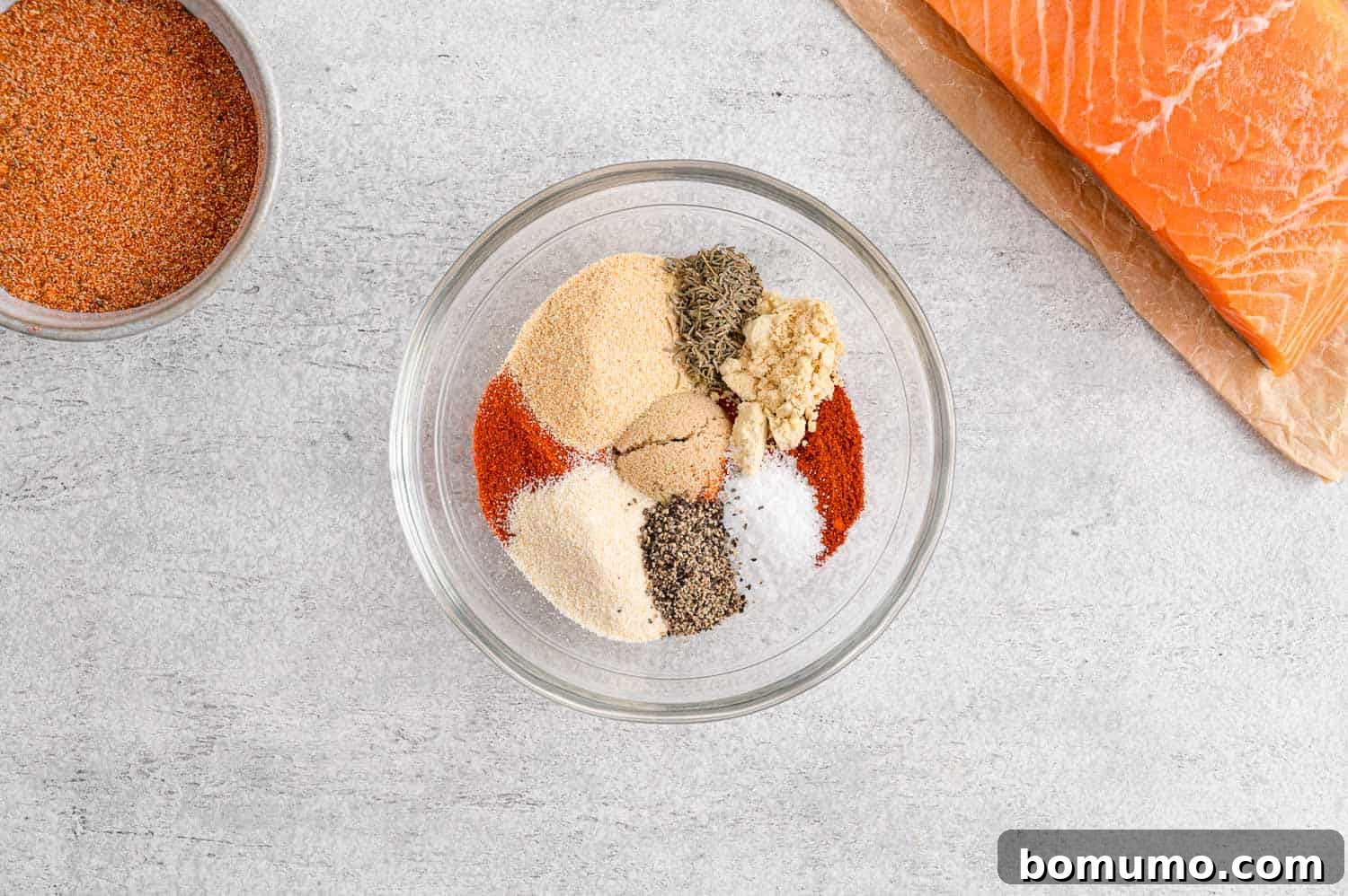 Individual spices for the salmon seasoning are poured into a small glass bowl, not yet mixed, ready for blending.