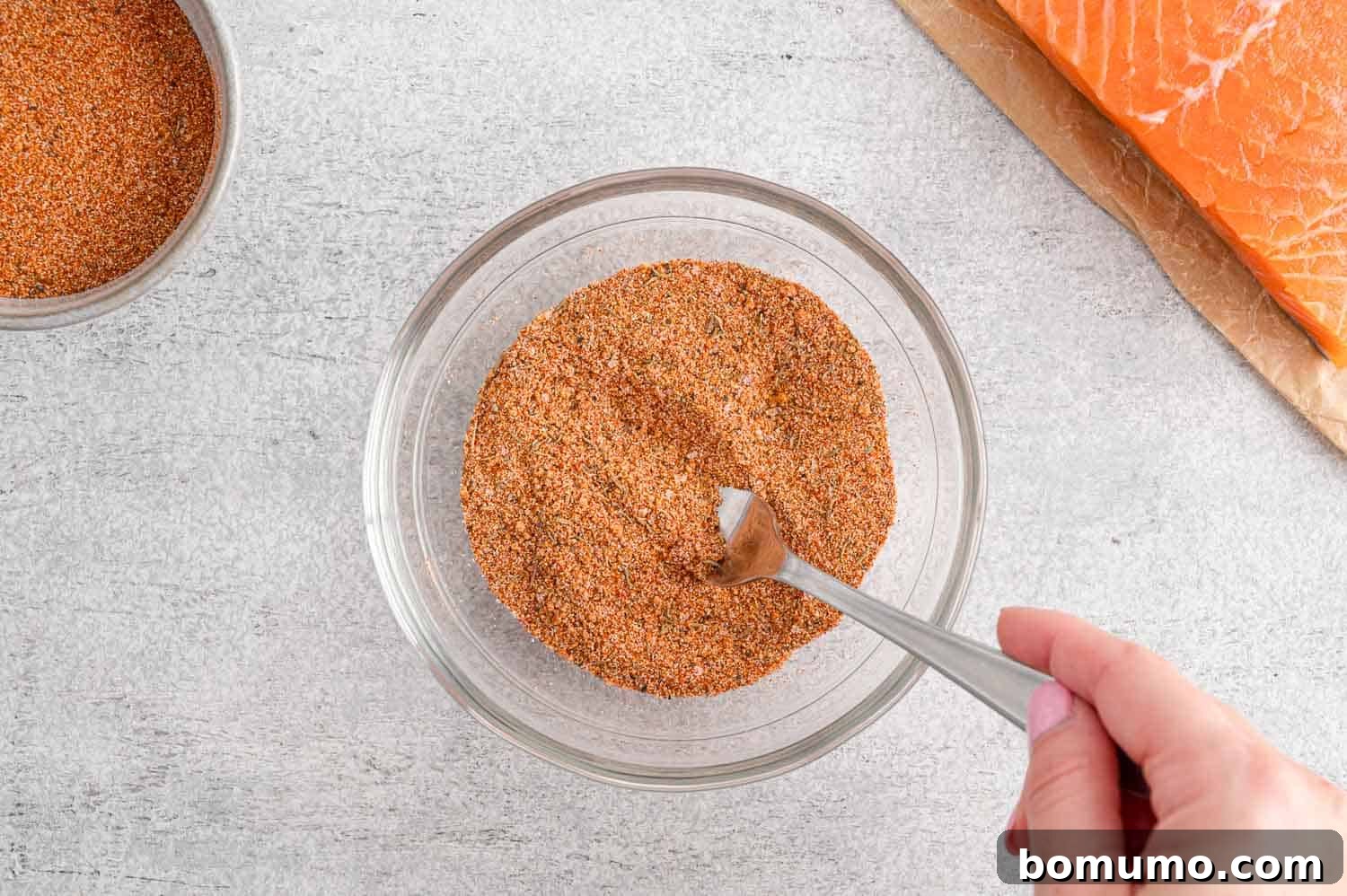 The spices for the salmon seasoning are being expertly mixed together with a small spoon in a glass bowl, ensuring an even blend.