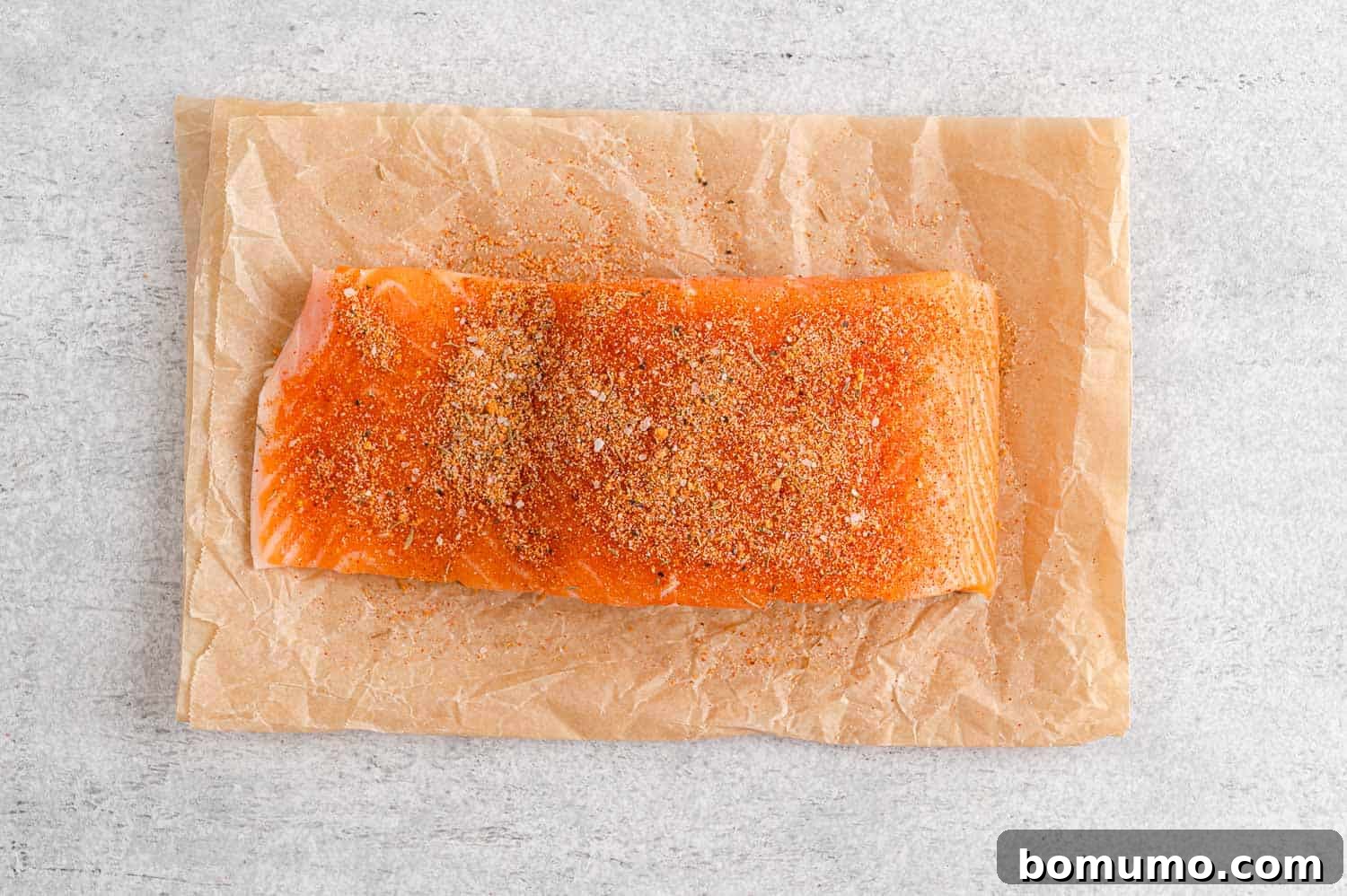 A raw salmon fillet, meticulously placed on parchment paper, is generously sprinkled with the vibrant salmon seasoning, ready for cooking.