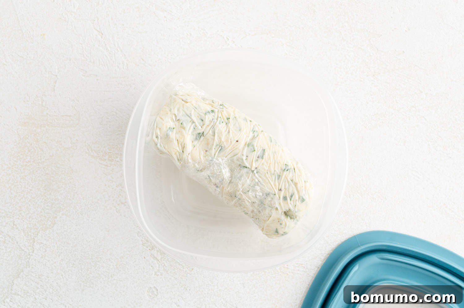 Log of garlic butter wrapped in plastic, placed inside a storage container for freezing.
