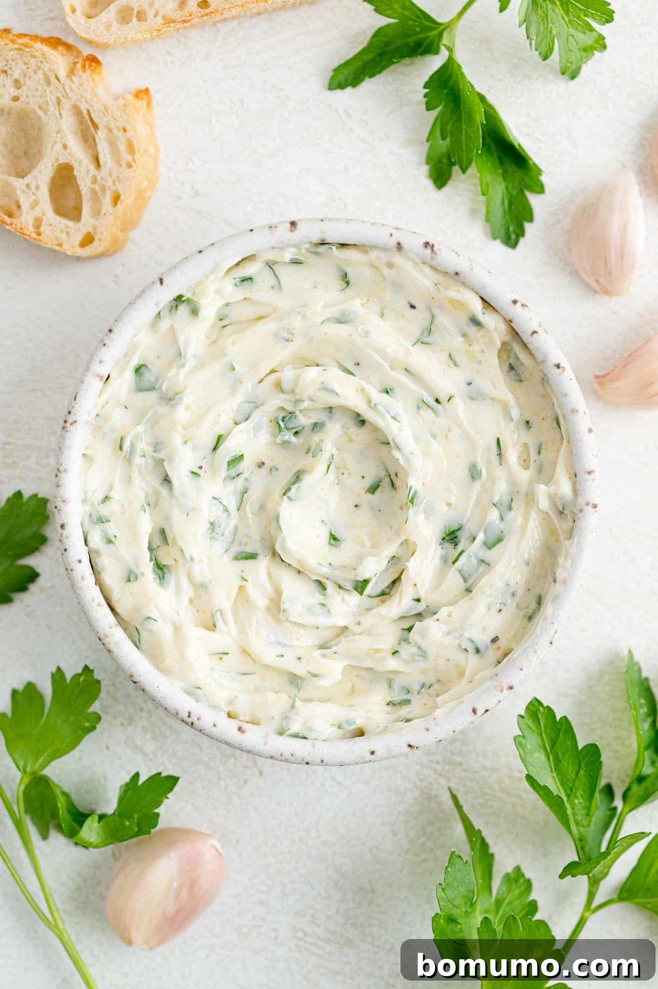 A bowl of garlic butter surrounded by fresh ingredients like garlic cloves and parsley, ready for preparation.