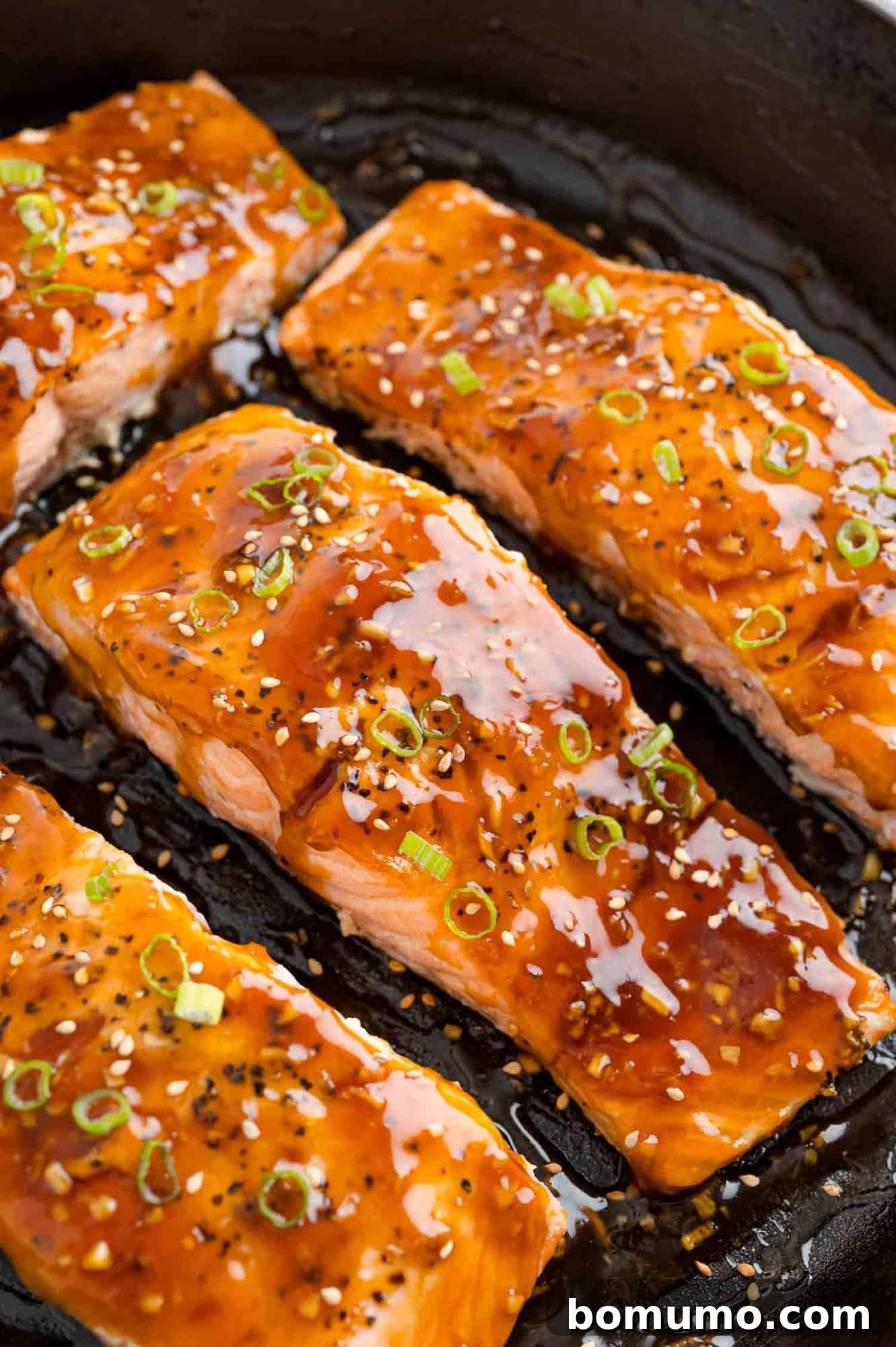 Golden brown teriyaki salmon fillets garnished with sesame seeds and chopped green onions in a sleek black frying pan.