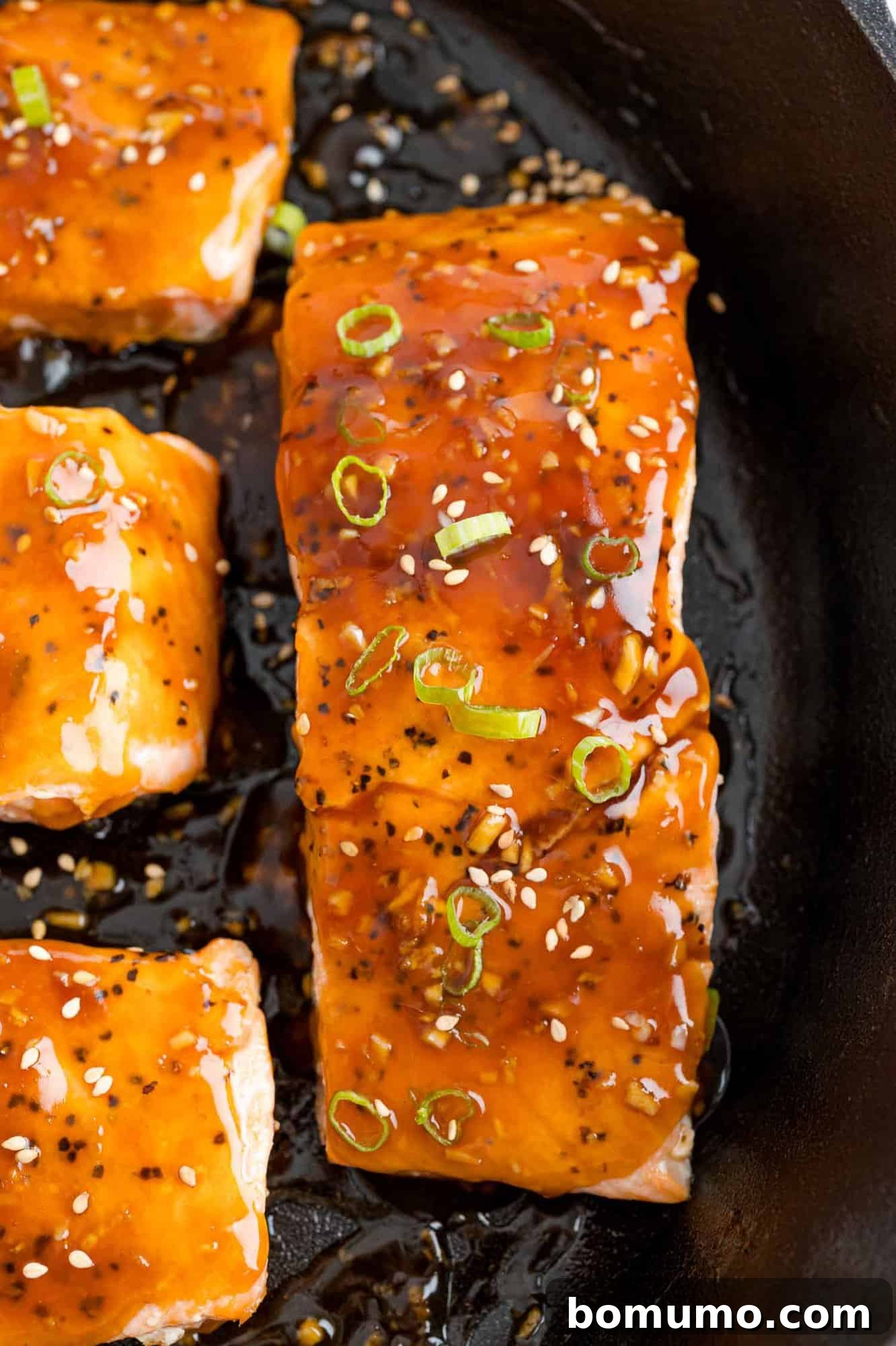 A close-up view of a perfectly cooked teriyaki salmon fillet, showcasing its glistening glaze, sesame seeds, and green onion garnish.