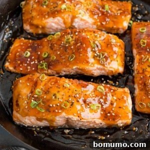 Teriyaki salmon fillets glistening in a black frying pan, ready to be served.