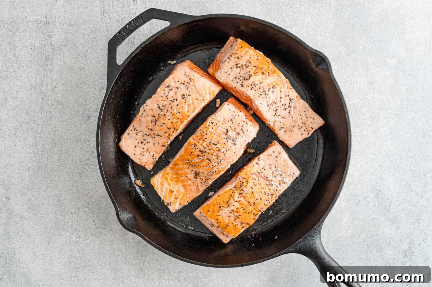Expertly seared salmon fillets, lightly browned and ready for the oven, resting skin-side down in a frying pan.