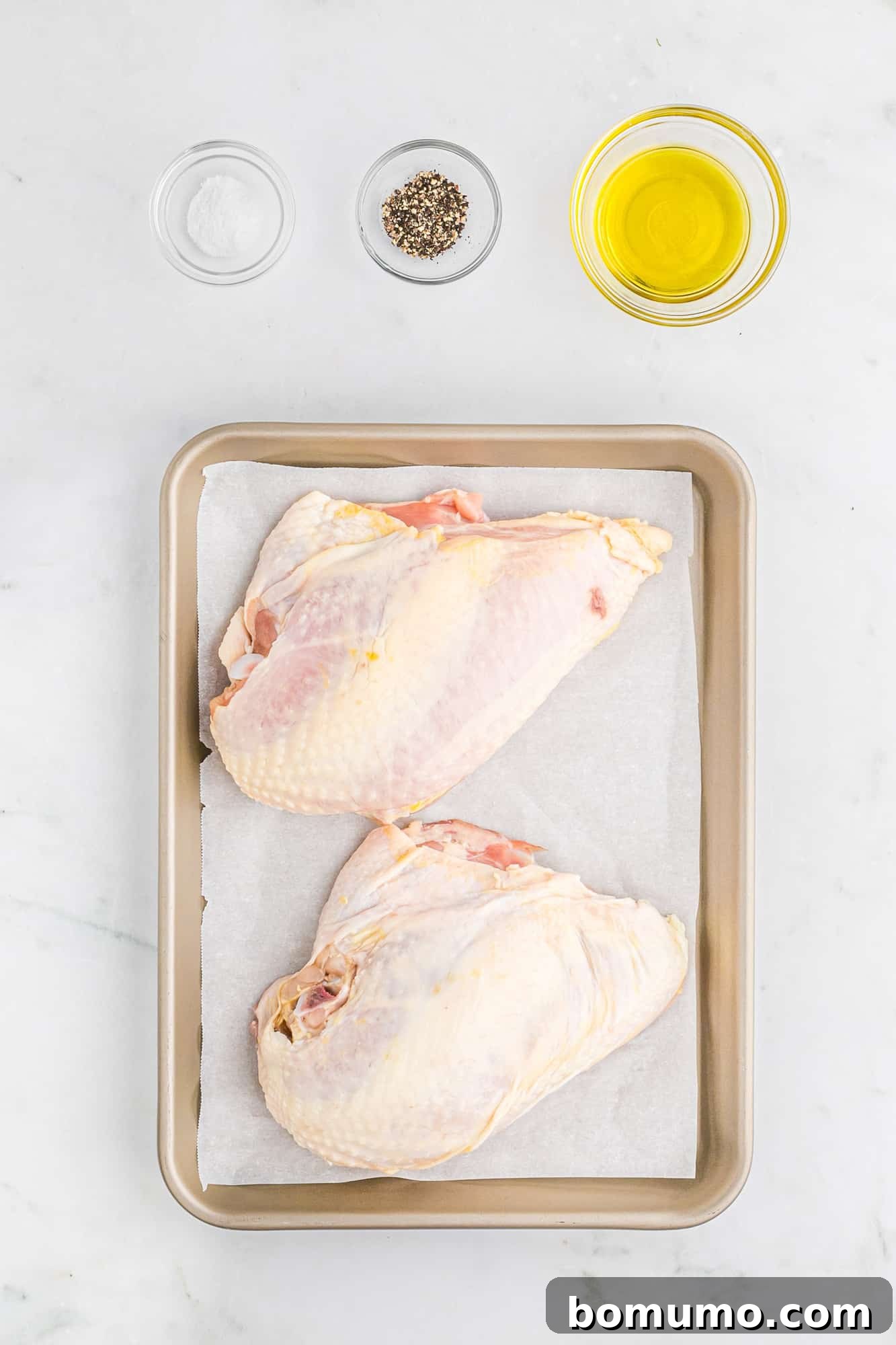 Fresh ingredients laid out on a counter for roasting chicken breasts: split chicken breasts, olive oil, salt, and pepper.