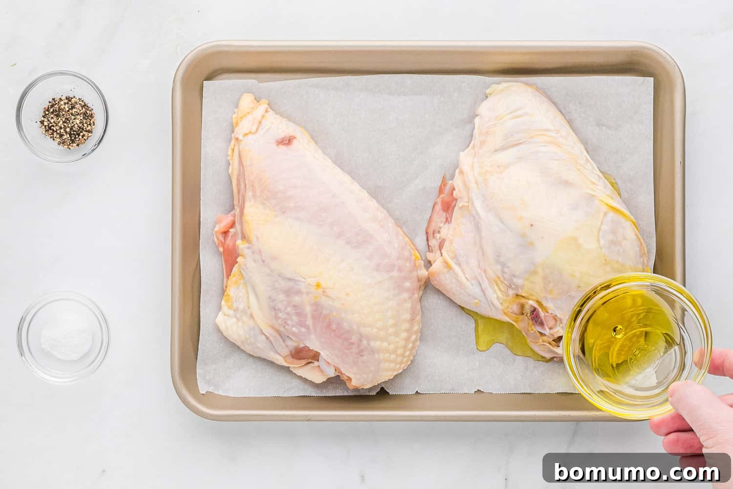 Olive oil being drizzled over raw, bone-in chicken breasts on a baking sheet, preparing them for roasting.