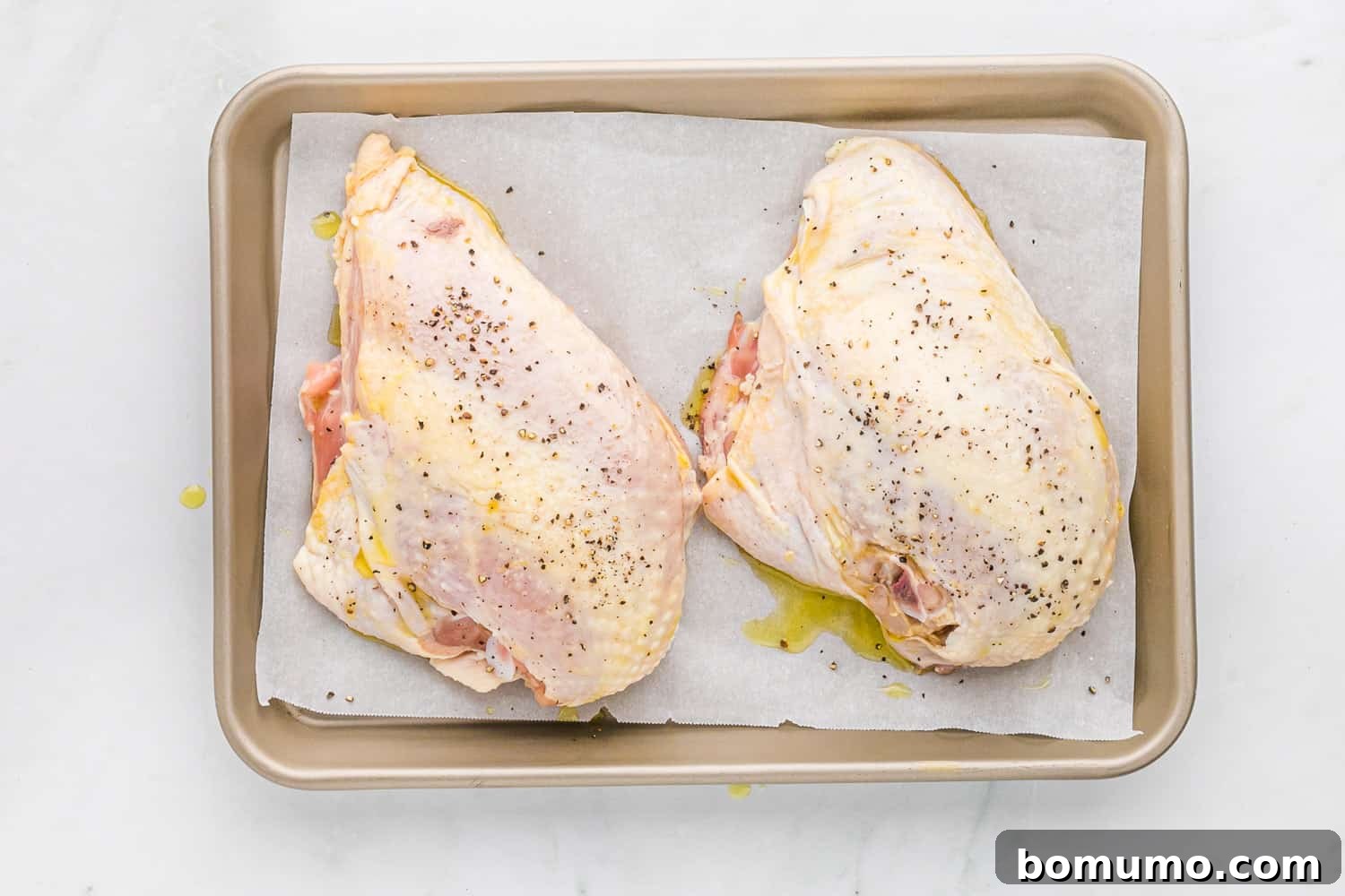 Raw bone-in chicken breasts seasoned with salt and pepper on a baking sheet, ready for the oven.