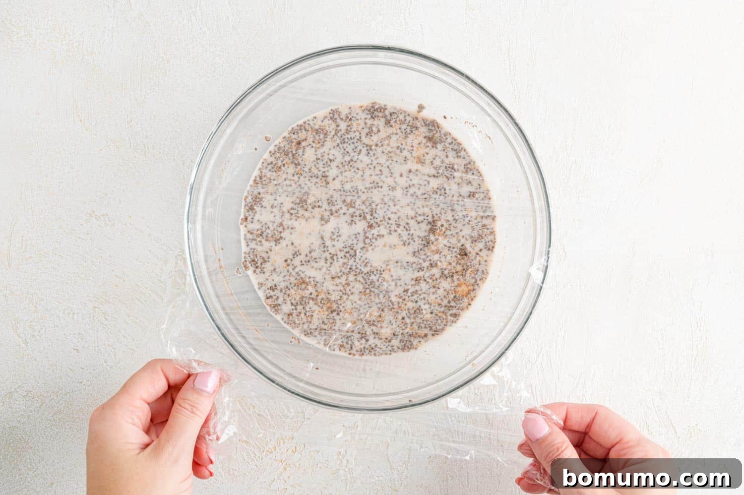 A glass bowl of unchilled chia pudding being covered with plastic wrap.