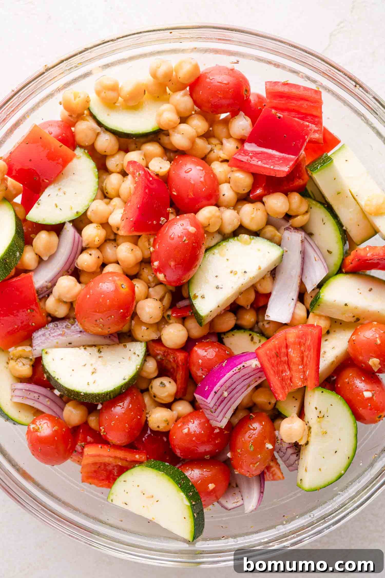 A glass bowl filled with a chickpeas, cherry tomatoes, cucumber slices, red bell pepper, and red onion, seasoned with herbs.