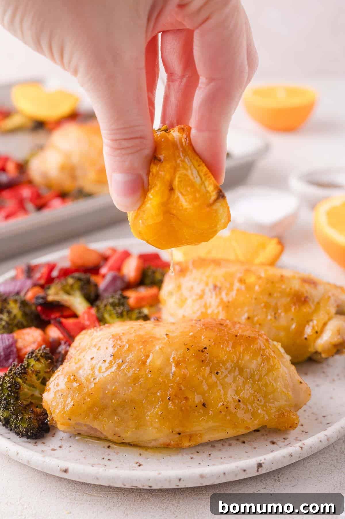 A hand squeezes an orange slice over roasted chicken thighs and vegetables on a plate, with more food and orange halves in the background.