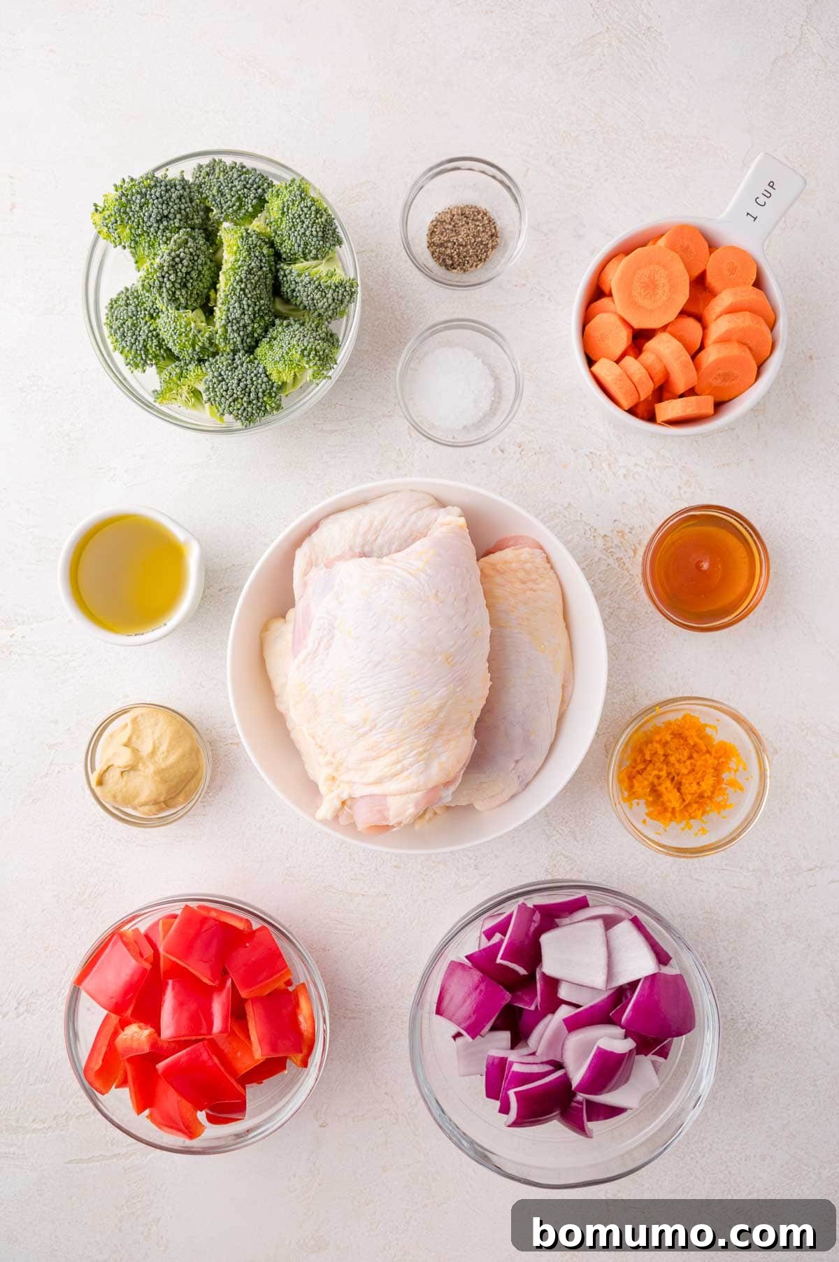 Ingredients for Sheet Pan Citrus Chicken and Rainbow Vegetables arranged in bowls on a white surface: raw chicken thighs, broccoli, carrots, red bell pepper, red onion, olive oil, mustard, honey, orange zest, salt, and pepper.