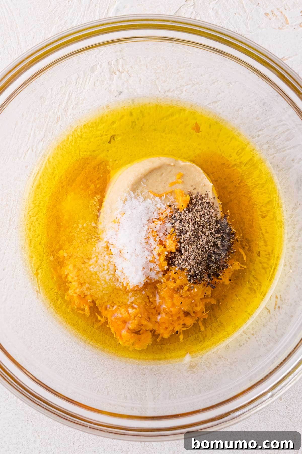 A glass bowl containing olive oil, mustard, salt, black pepper, and citrus zest on a light-colored surface.
