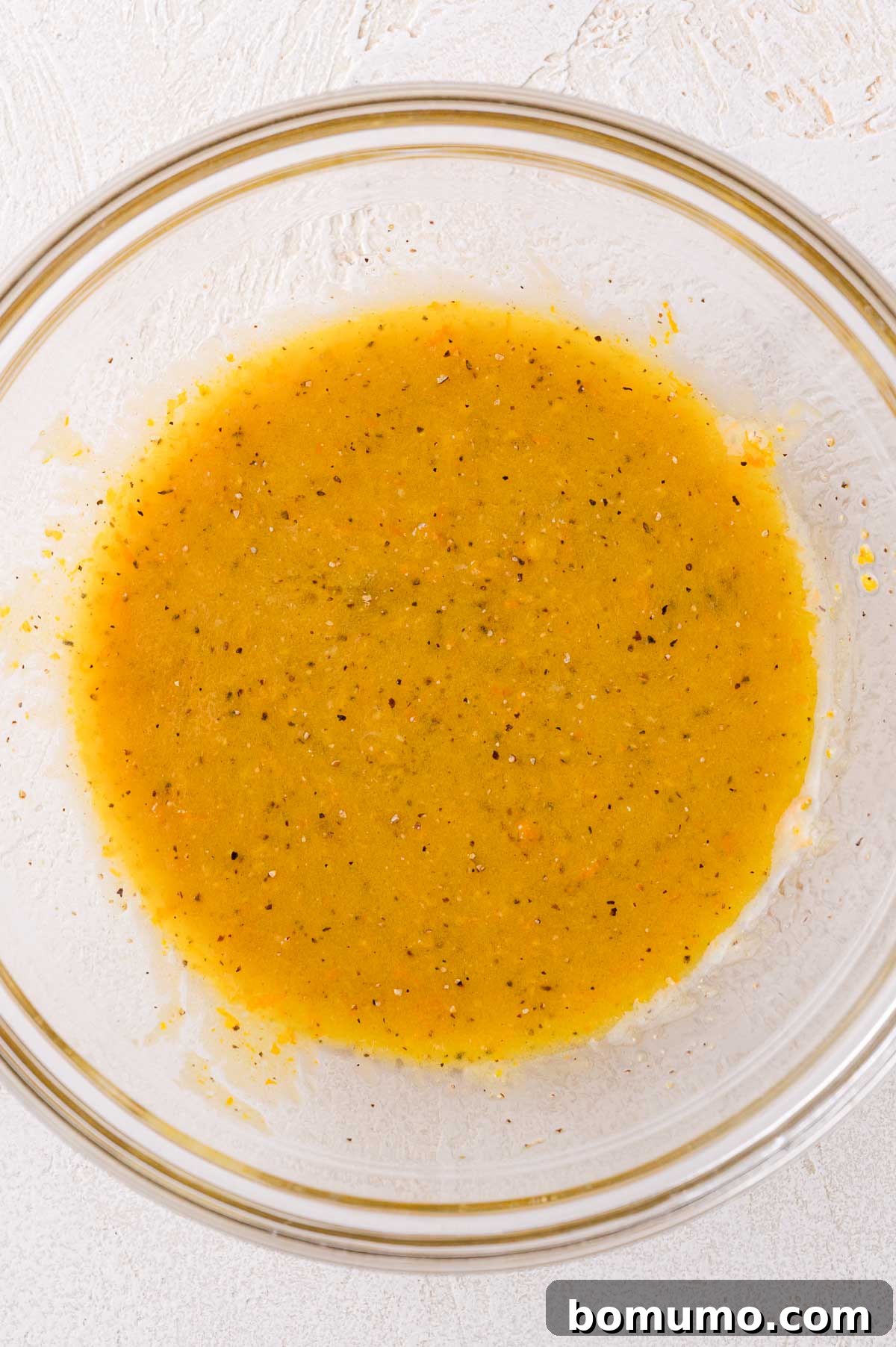 A glass bowl containing a yellow, speckled liquid mixture (citrus honey mustard sauce) on a light textured surface.