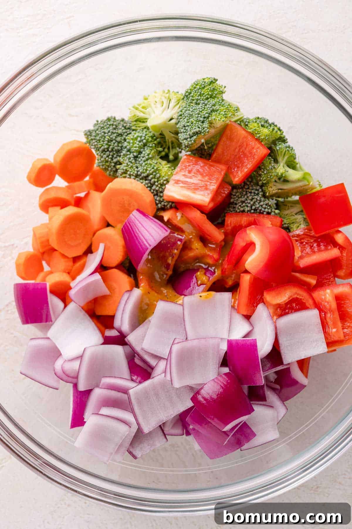 A glass bowl filled with chopped red onions, carrots, red bell peppers, and broccoli florets on a light surface.