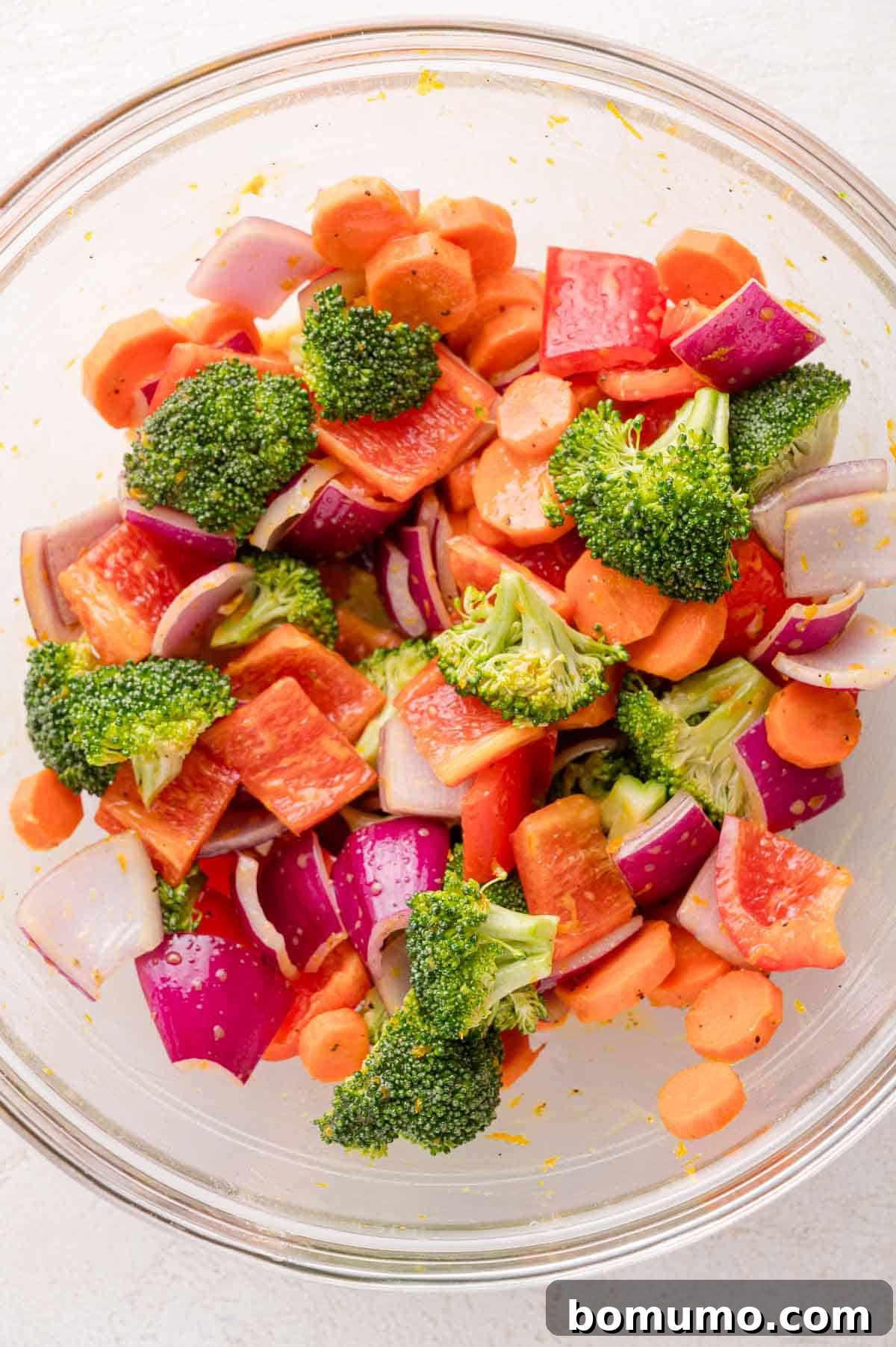 A glass bowl filled with chopped broccoli, red bell pepper, red onion, and carrots.