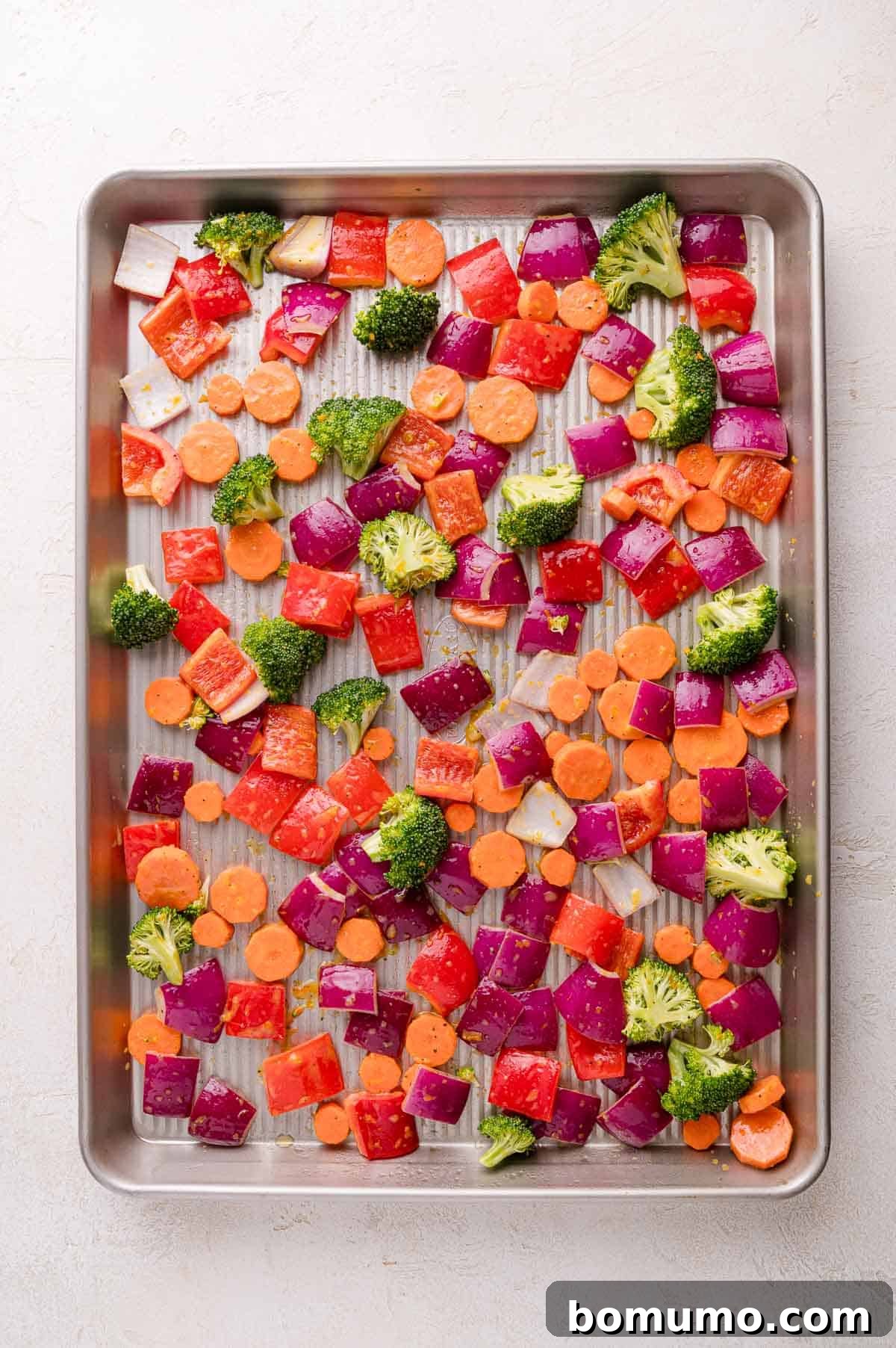 Chopped red onions, carrots, red bell peppers, and broccoli florets spread out on a metal baking sheet, ready for roasting.