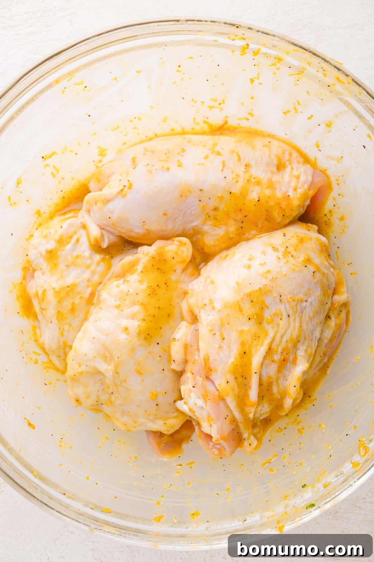 Raw chicken breasts coated in a yellow marinade with spices in a clear glass bowl on a white surface.
