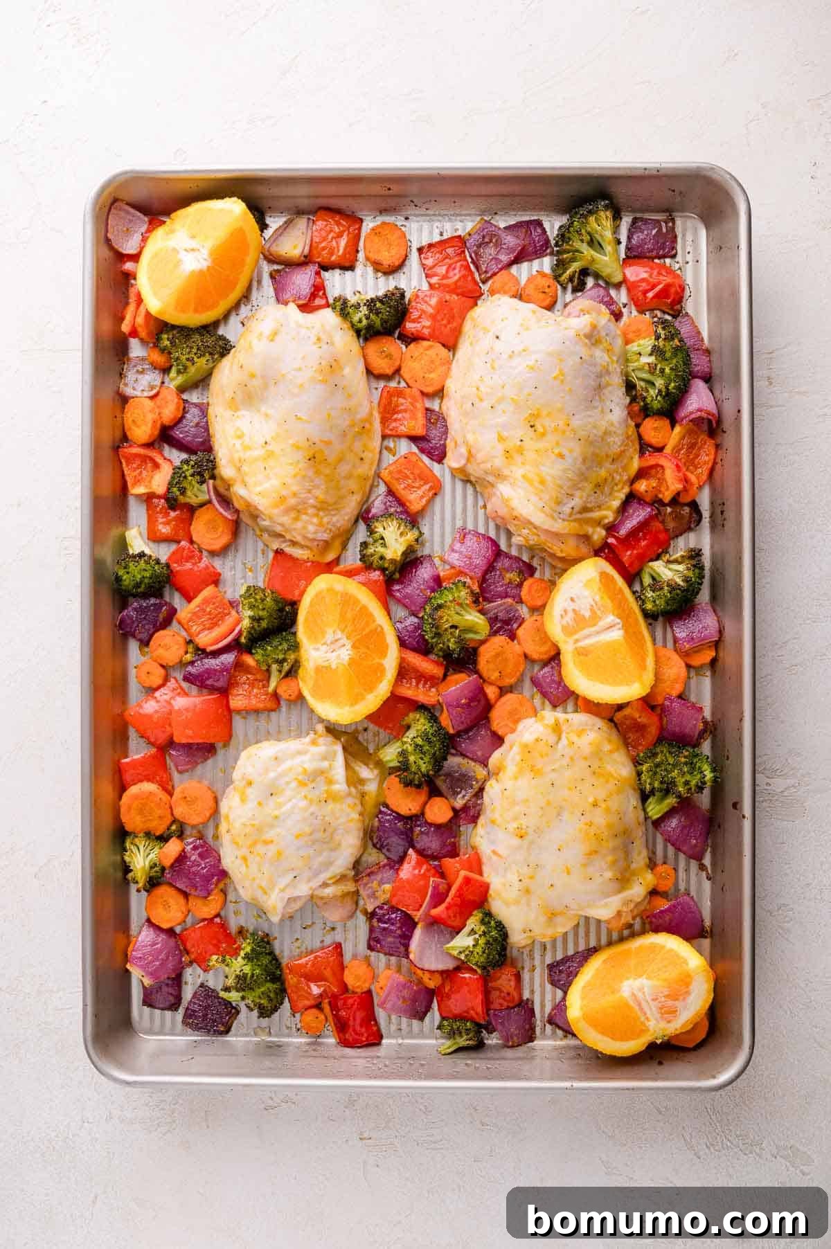 A sheet pan with raw chicken thighs, chopped carrots, red bell peppers, broccoli, and red onion, garnished with orange halves, ready to be cooked.