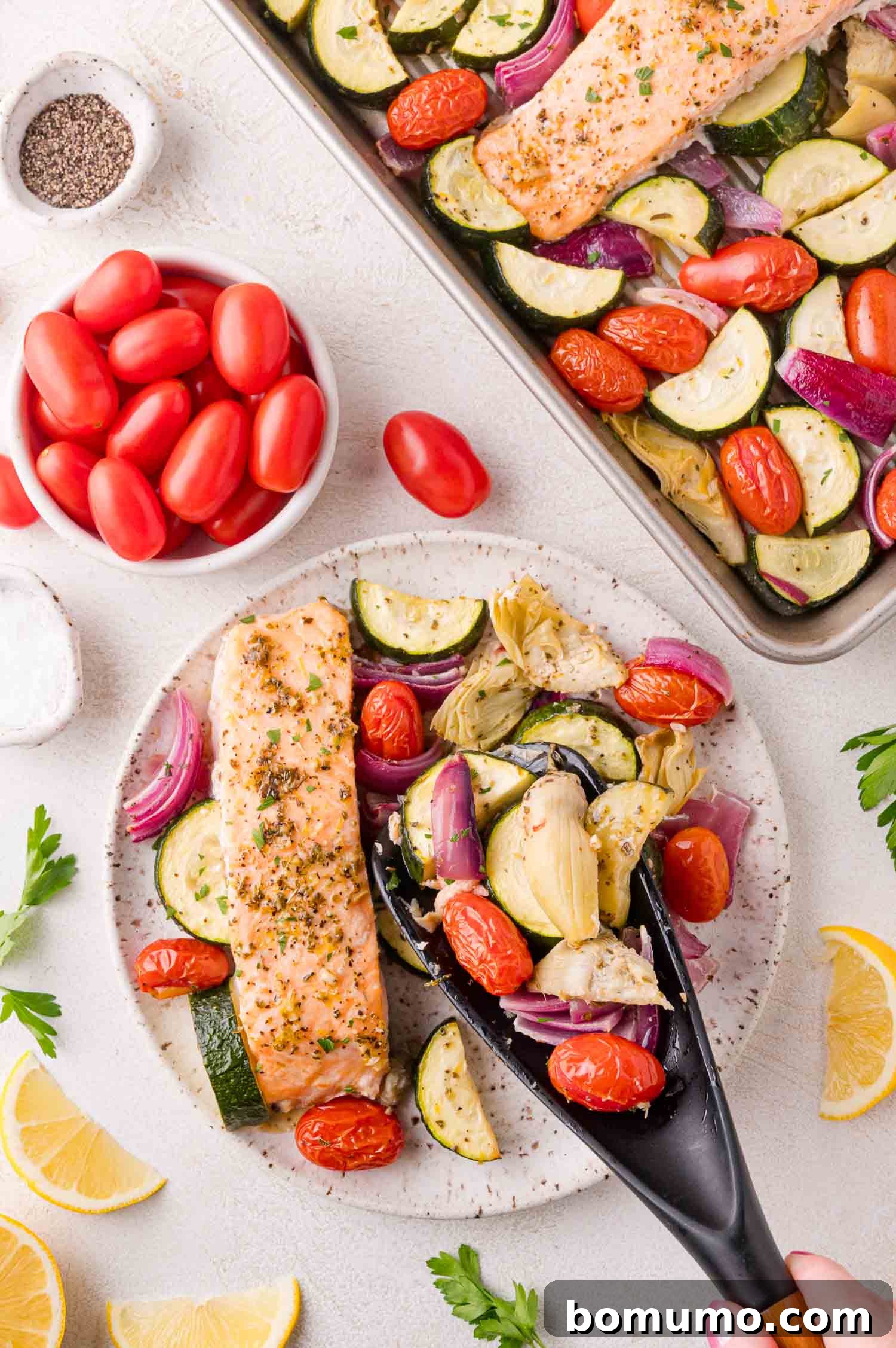 Greek Salmon Sheet Pan Dinner served on a plate with roasted zucchini, tomatoes, red onion, and artichokes. A sheet pan and bowl of tomatoes are in the background.