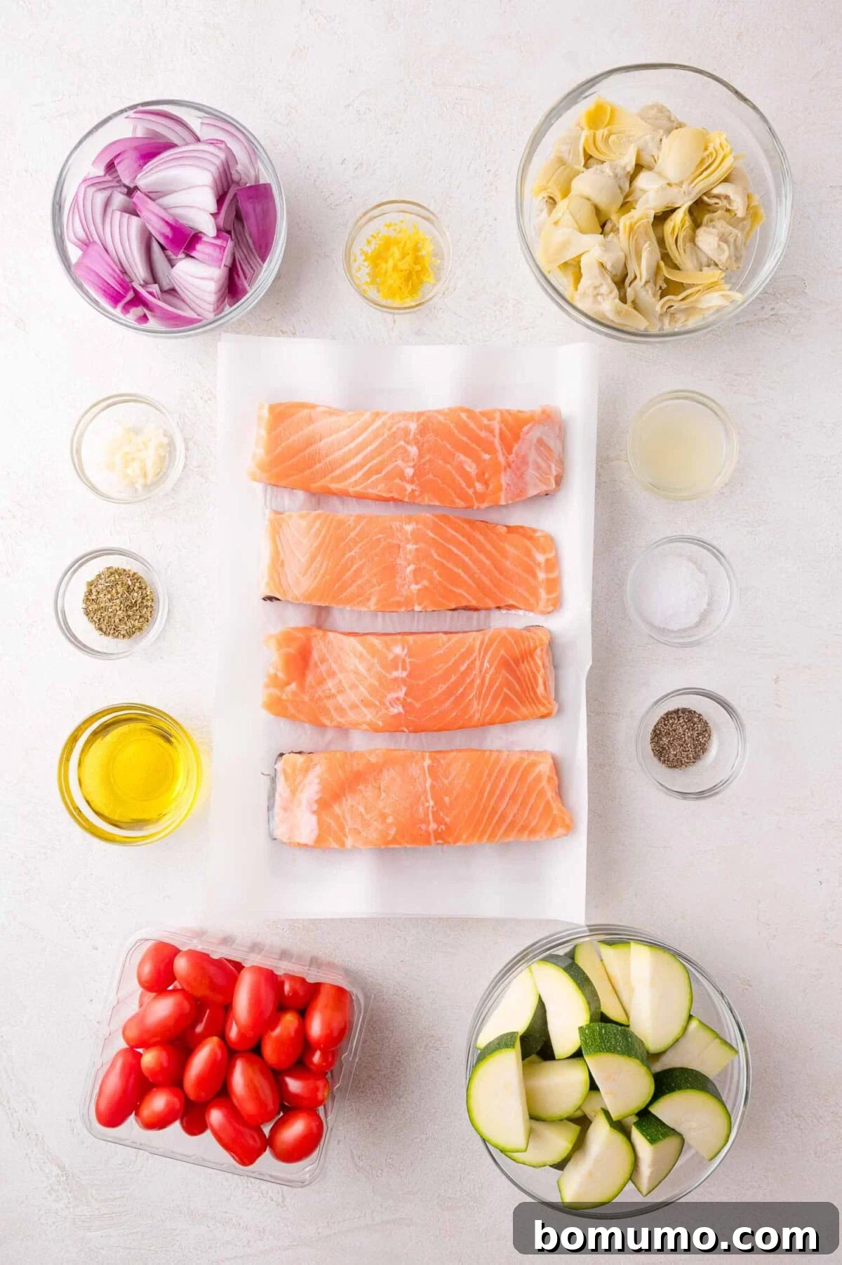Ingredients for Greek Salmon Sheet Pan Dinner: raw salmon fillets, red onion, artichoke hearts, zucchini, grape tomatoes, lemon zest, olive oil, seasoning, lemon juice, and garlic.