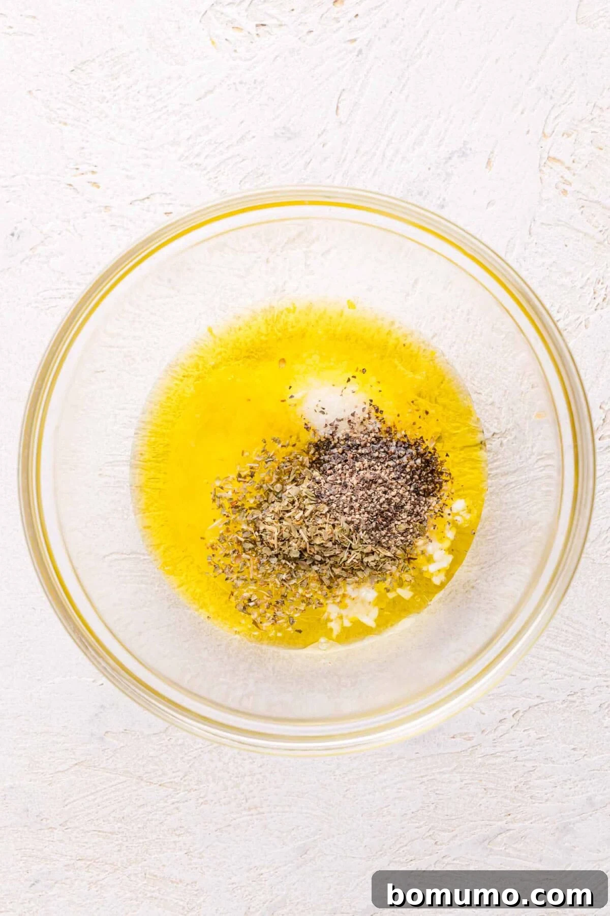 Olive oil, minced garlic, dried herbs, salt, and pepper in a glass bowl.