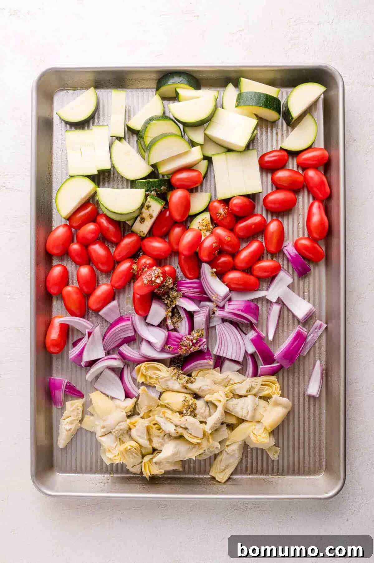 Chopped zucchini, grape tomatoes, red onion, and artichoke hearts arranged on a baking sheet.