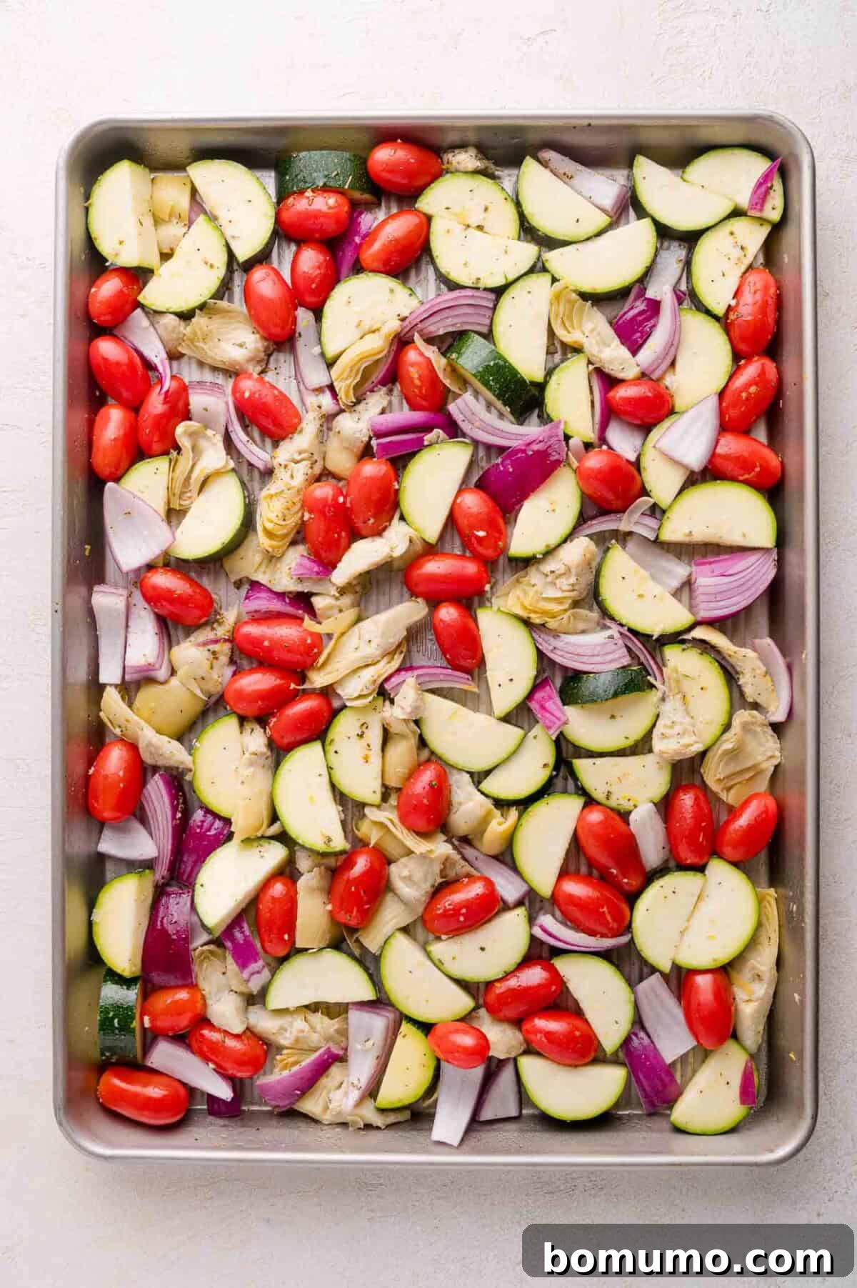Baking sheet with chopped and seasoned zucchini, red onion, grape tomatoes, and artichoke hearts.