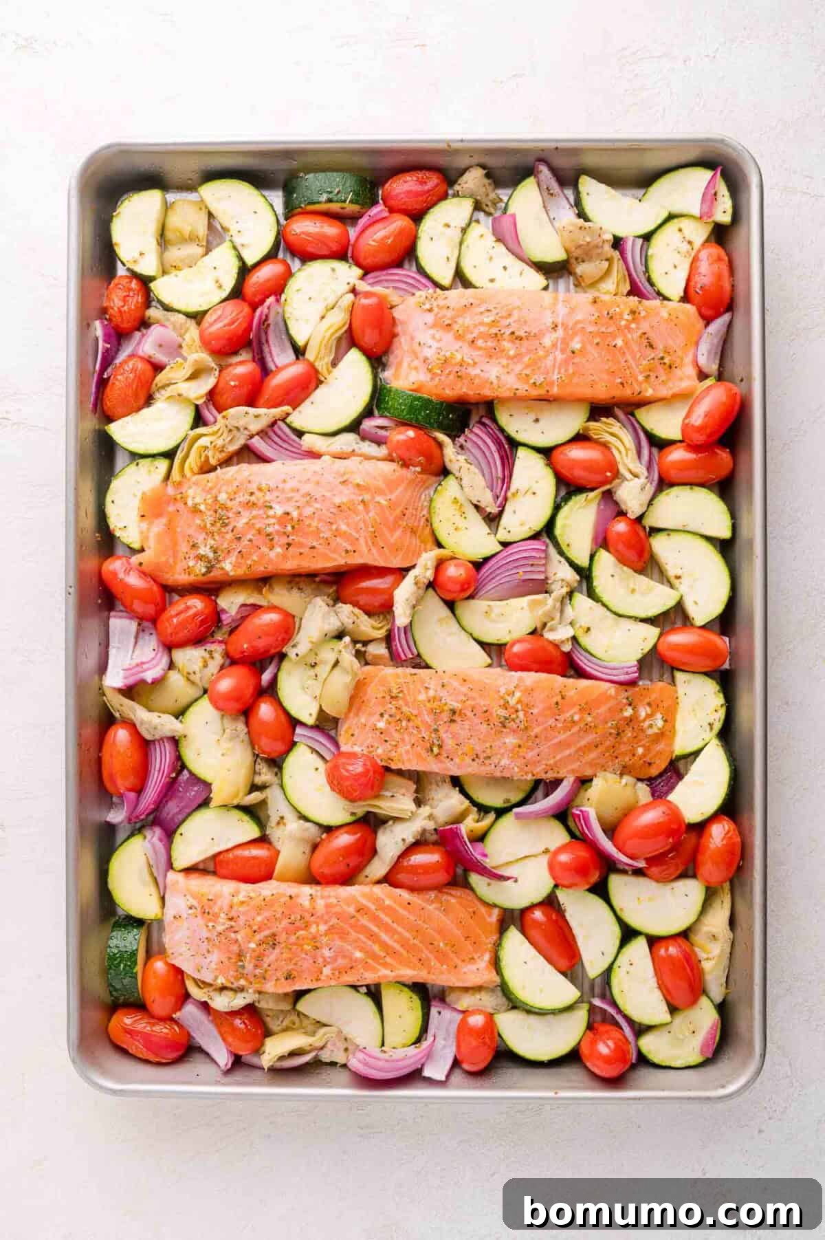 Raw salmon fillets on top of sliced zucchini, red onion, cherry tomatoes, and artichoke hearts on a baking sheet.