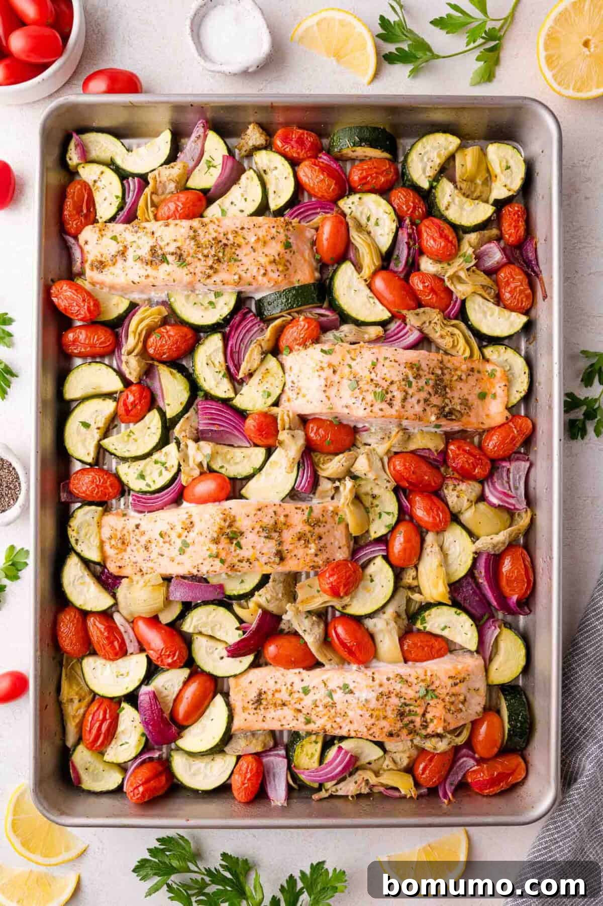 Salmon fillets surrounded by zucchini, cherry tomatoes, red onion, and artichoke hearts, seasoned and ready to roast.