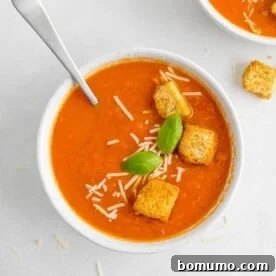 Tomato soup in white bowl garnished with croutons, cheese, basil.