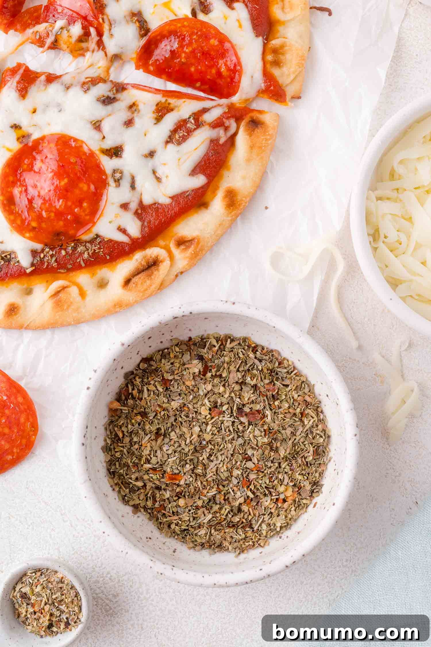 Pizza topped with cheese, bowl of pizza seasoning, and bowl of cheese