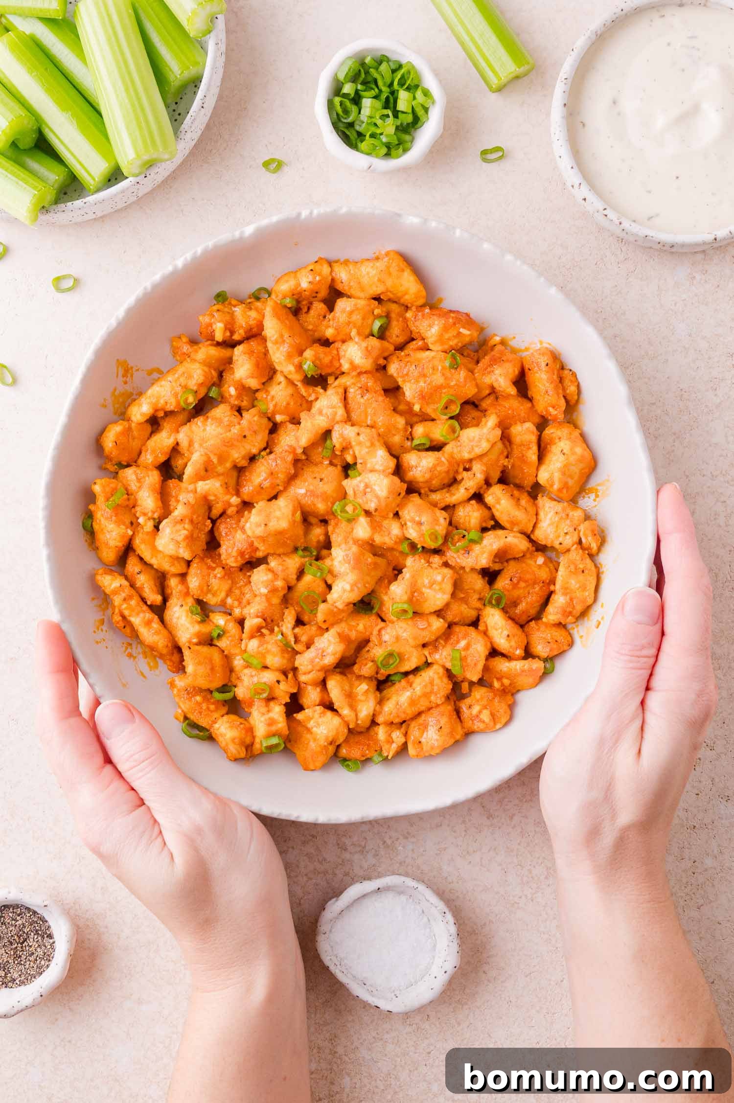 Buffalo Chicken Poppers 13 A bowl of buffalo chicken bites garnished with green onions, surrounded by celery sticks, ranch dressing, and seasonings, with two hands holding the bowl.
