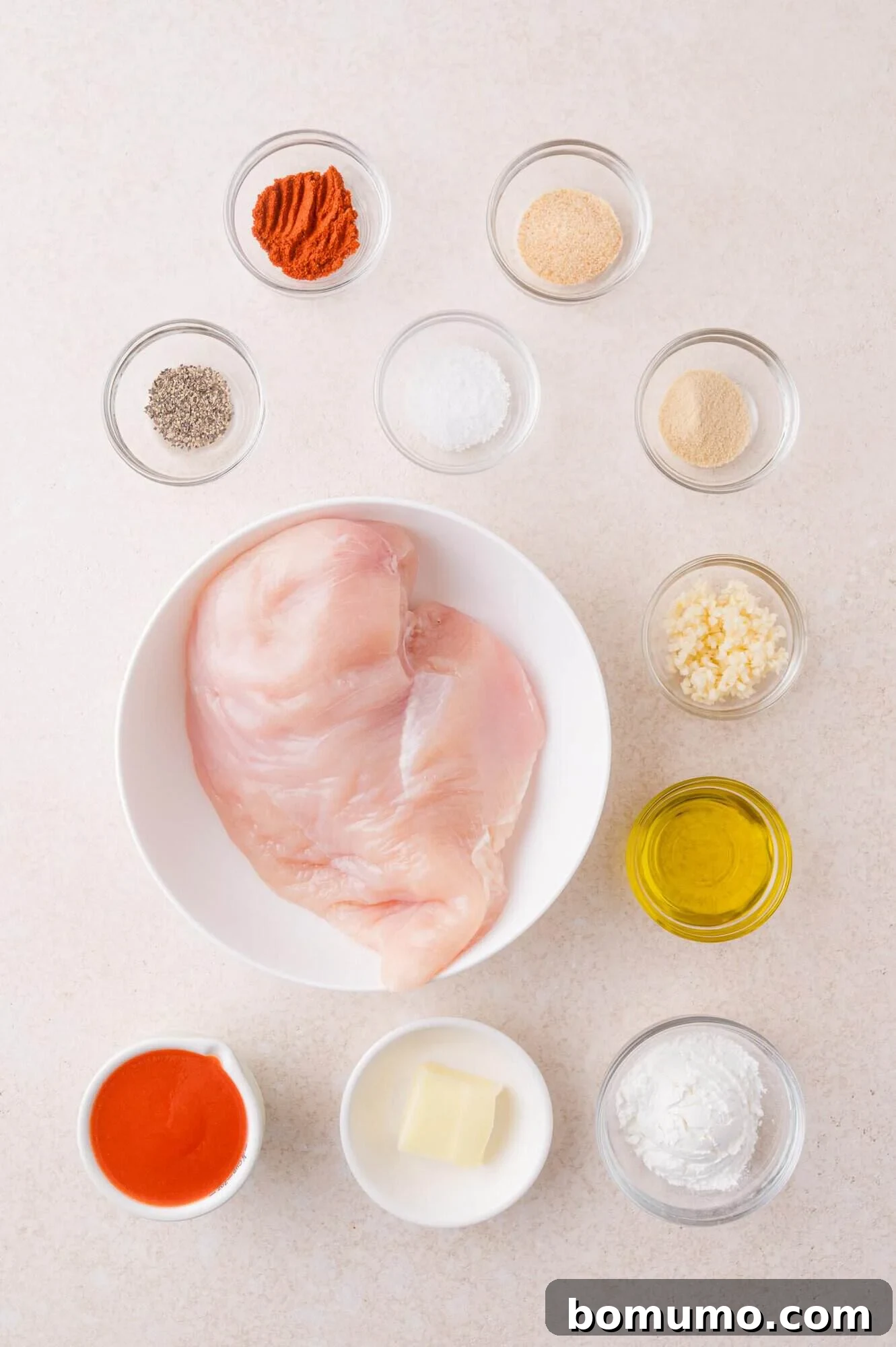 Buffalo Chicken Poppers 3 Raw chicken breast in a white bowl surrounded by small bowls of spices, garlic, oil, hot sauce, butter, and cornstarch on a light surface.