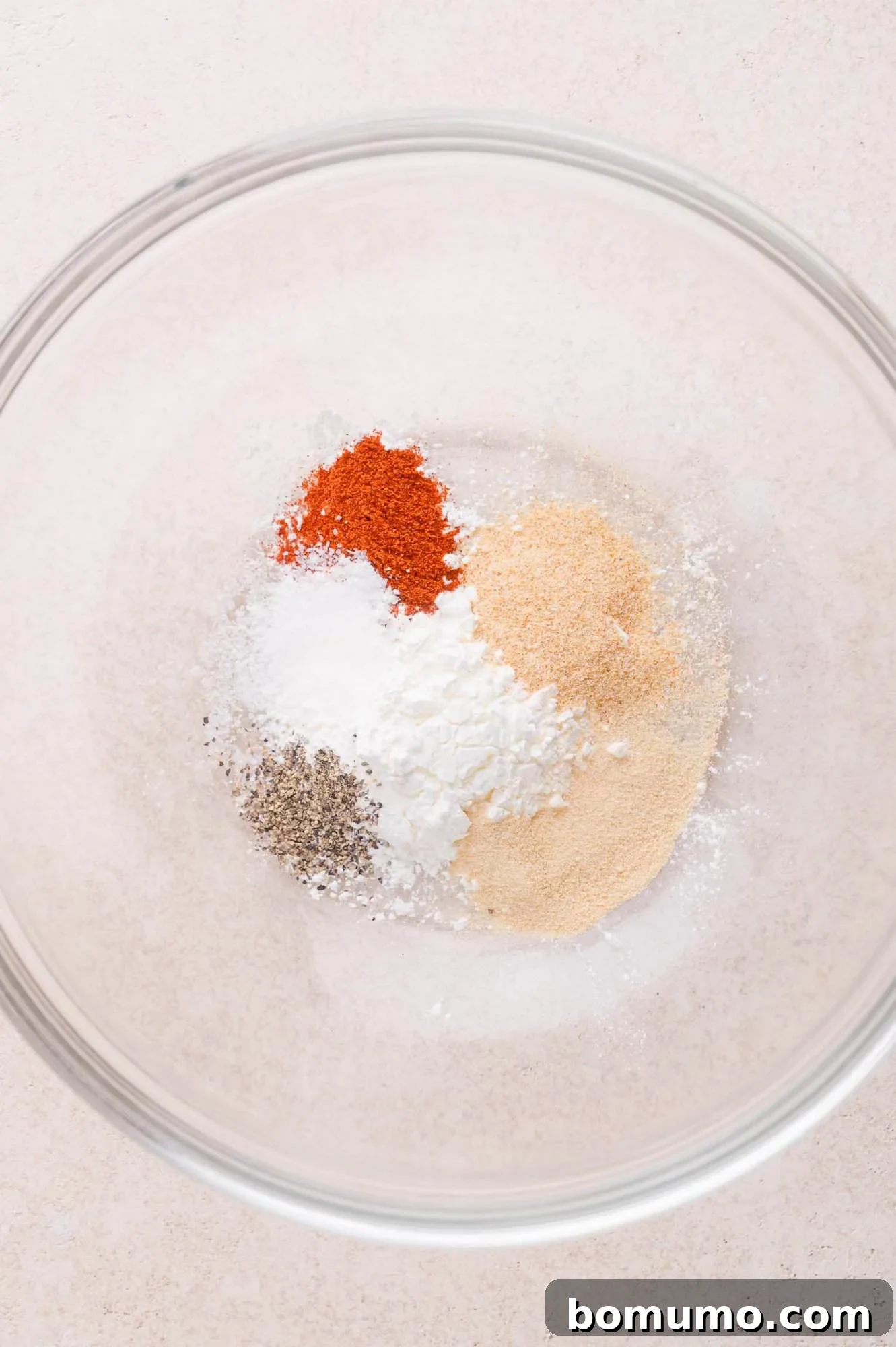 Buffalo Chicken Poppers 4 A clear glass bowl containing separate piles of paprika, black pepper, salt, cornstarch, garlic powder, and onion powder on a light surface.