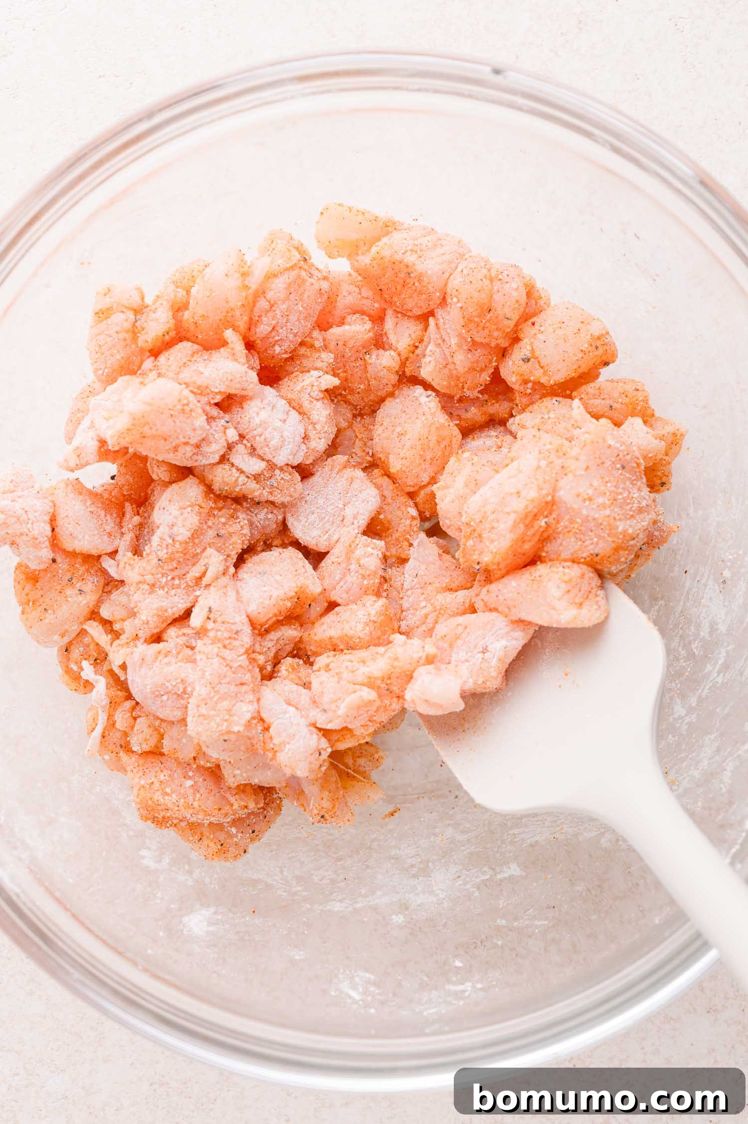 Buffalo Chicken Poppers 8 Diced raw chicken pieces coated with seasoning in a clear glass bowl, being mixed with a white spatula.