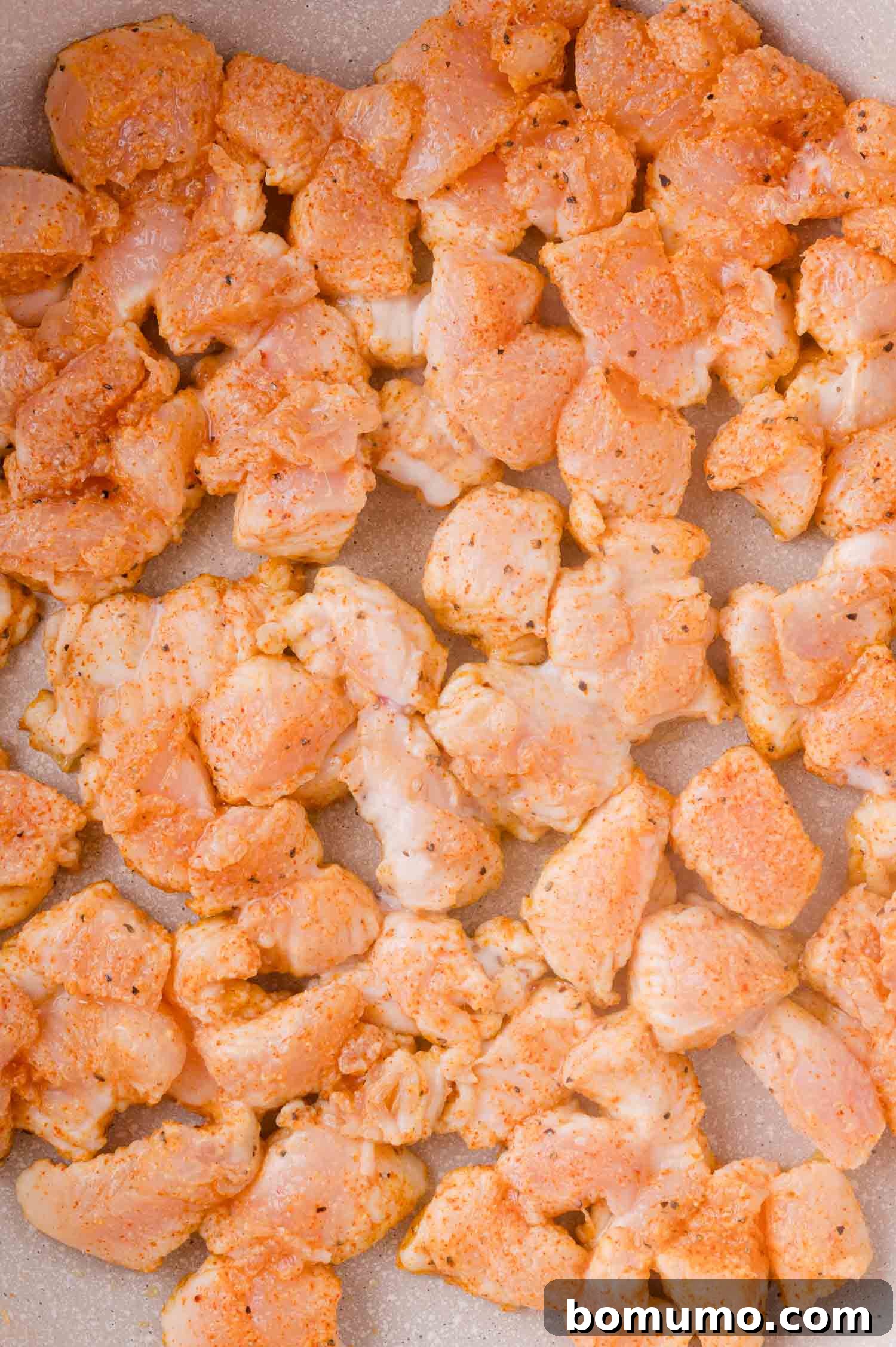 Buffalo Chicken Poppers 9 Raw, diced chicken breast coated with spices spread out in a single layer on a light-colored surface, ready to be cooked.