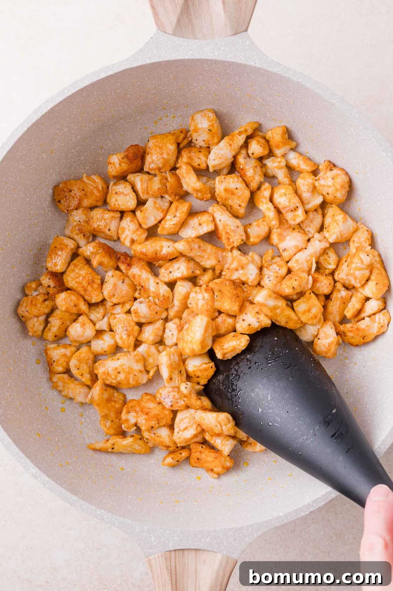 Buffalo Chicken Poppers 10 Diced, seasoned chicken cooking in a light-colored skillet is being stirred with a black spatula.