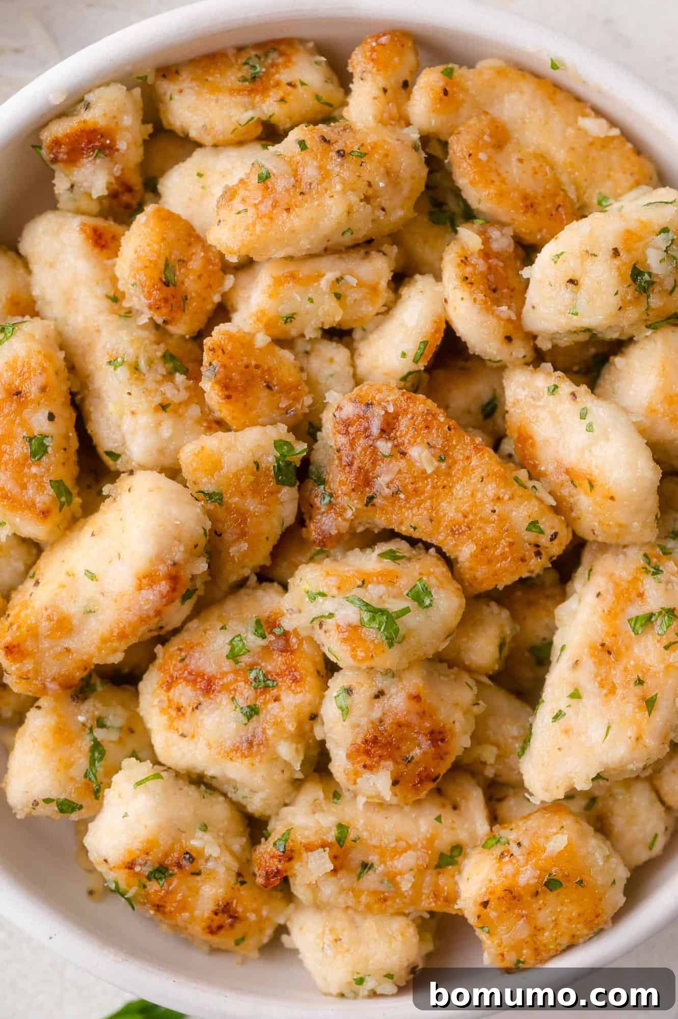 A bowl filled with golden-brown, pan-seared pieces of chicken garnished with parmesan and chopped herbs.