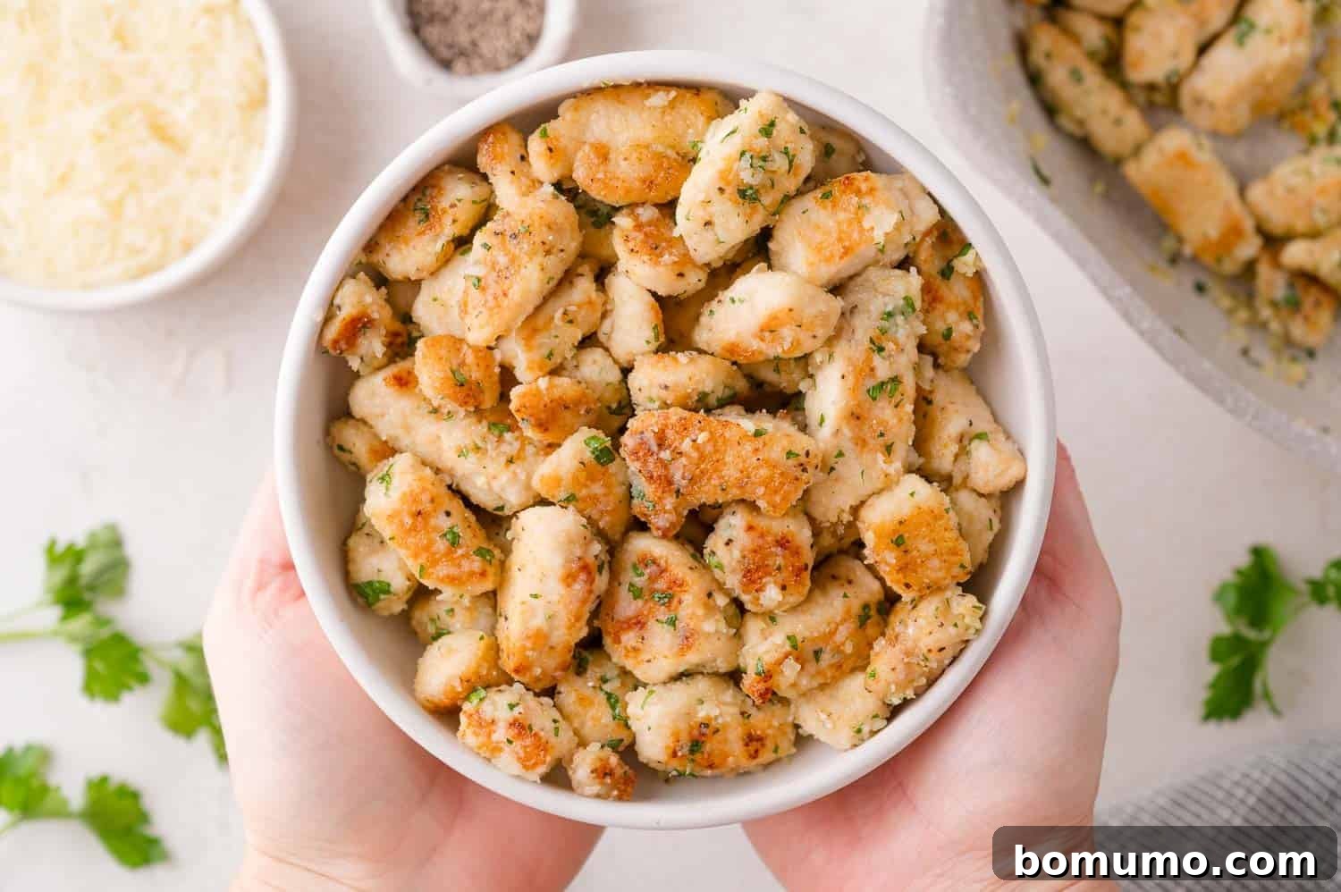 A bowl filled with cooked, golden-brown, bite-sized pieces of chicken garnished with herbs, held by two hands; grated cheese, pepper, and parsley are nearby.