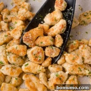 Sautéed, golden-brown chicken pieces garnished with garlic and herbs, being stirred with a black spatula in a pan.