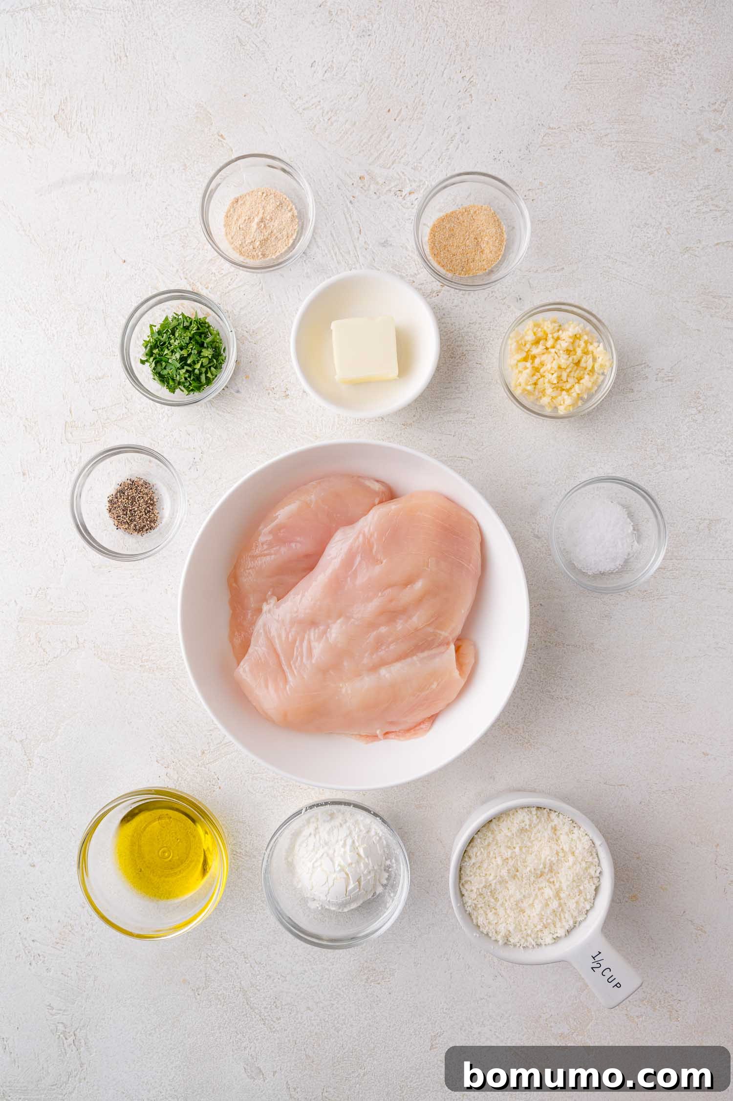 A bowl of raw chicken breasts surrounded by small bowls containing olive oil, butter, chopped herbs, garlic, grated cheese, and seasonings on a white surface.